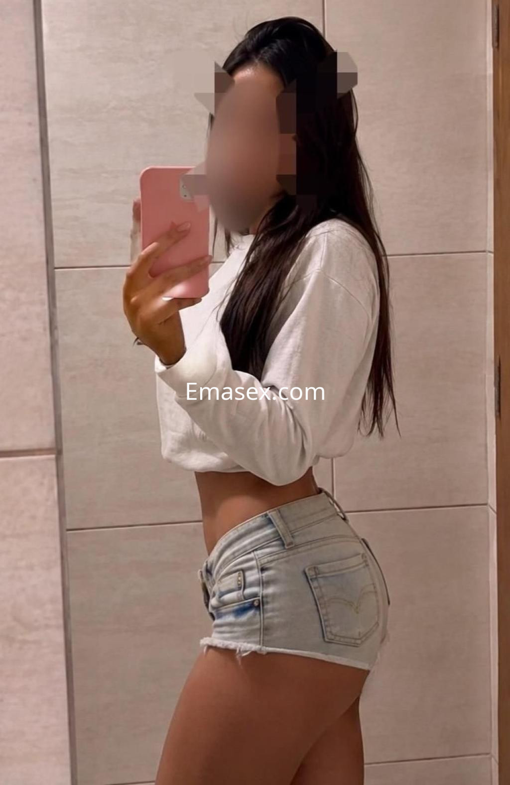 Puta con cabello largo y lacio, viste una camiseta blanca y shorts de mezclilla, posando en un baño en Málaga.