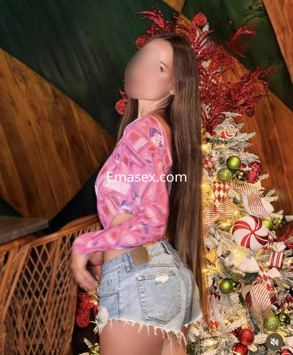 La puta en Valencia lleva una blusa de manga larga rosa y shorts de mezclilla, posando junto a un árbol navideño.