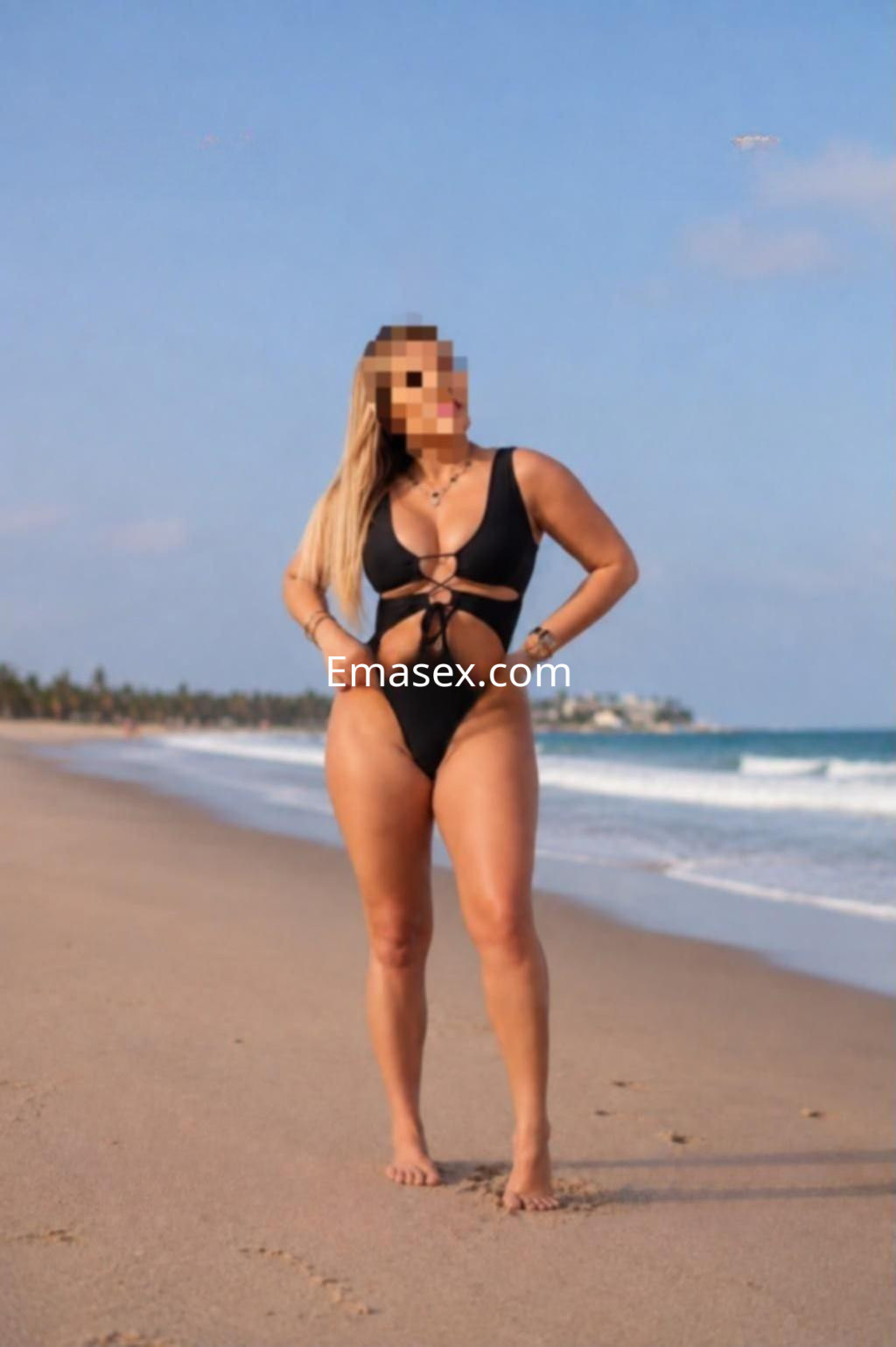Escort de Valencia posando en la playa con un traje de baño negro, cabello largo y rubio, mostrando confianza.