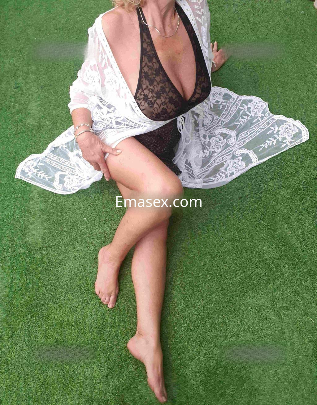 Escort sentada sobre césped artificial, lleva un atuendo negro con una chaqueta blanca de encaje en Madrid.