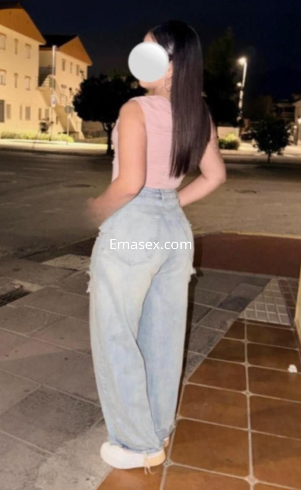 Escort en Madrid con blusa rosa y jeans desgastados, de espaldas en una calle iluminada por la noche.