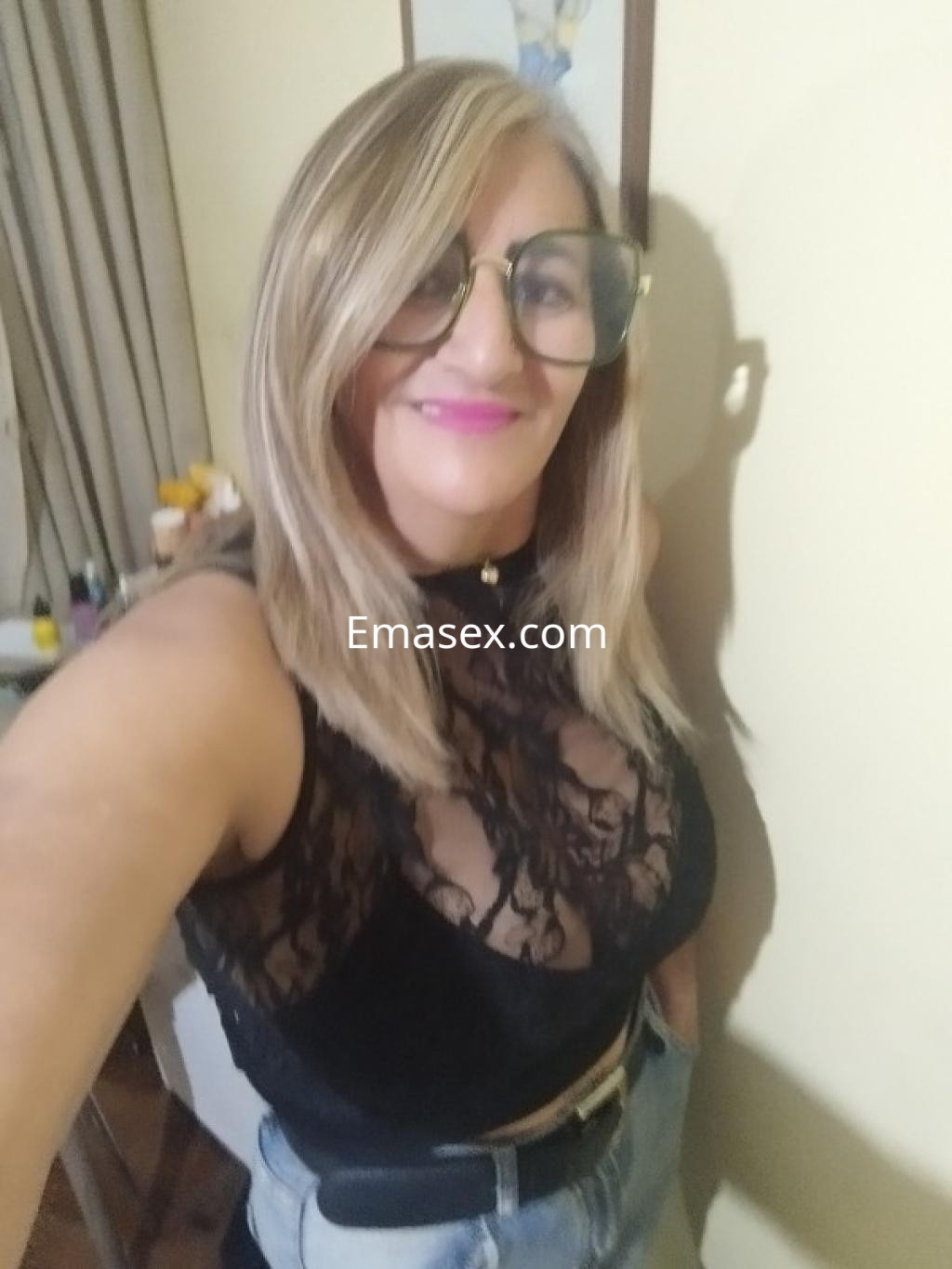 Escort en Coruña, con blusa de encaje negra y gafas grandes, sonríe frente a un fondo de pared amarilla.