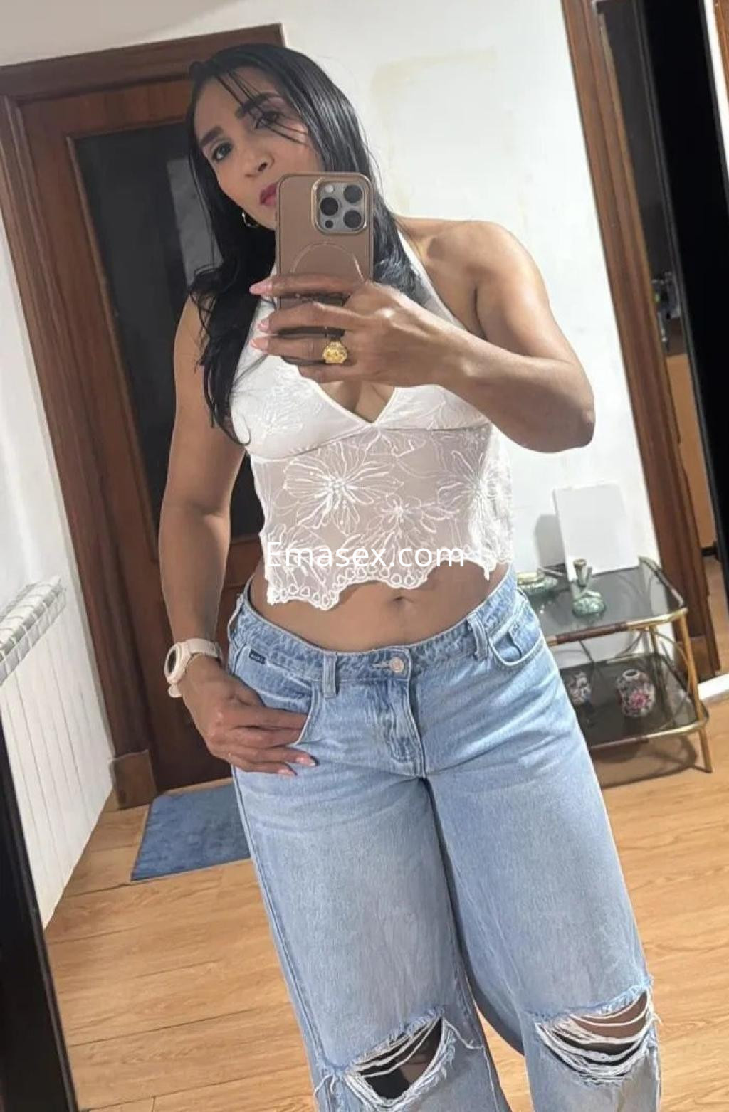 Escort en Burgos viste una blusa blanca de encaje y jeans rotos, posando frente a un espejo en un ambiente acogedor.