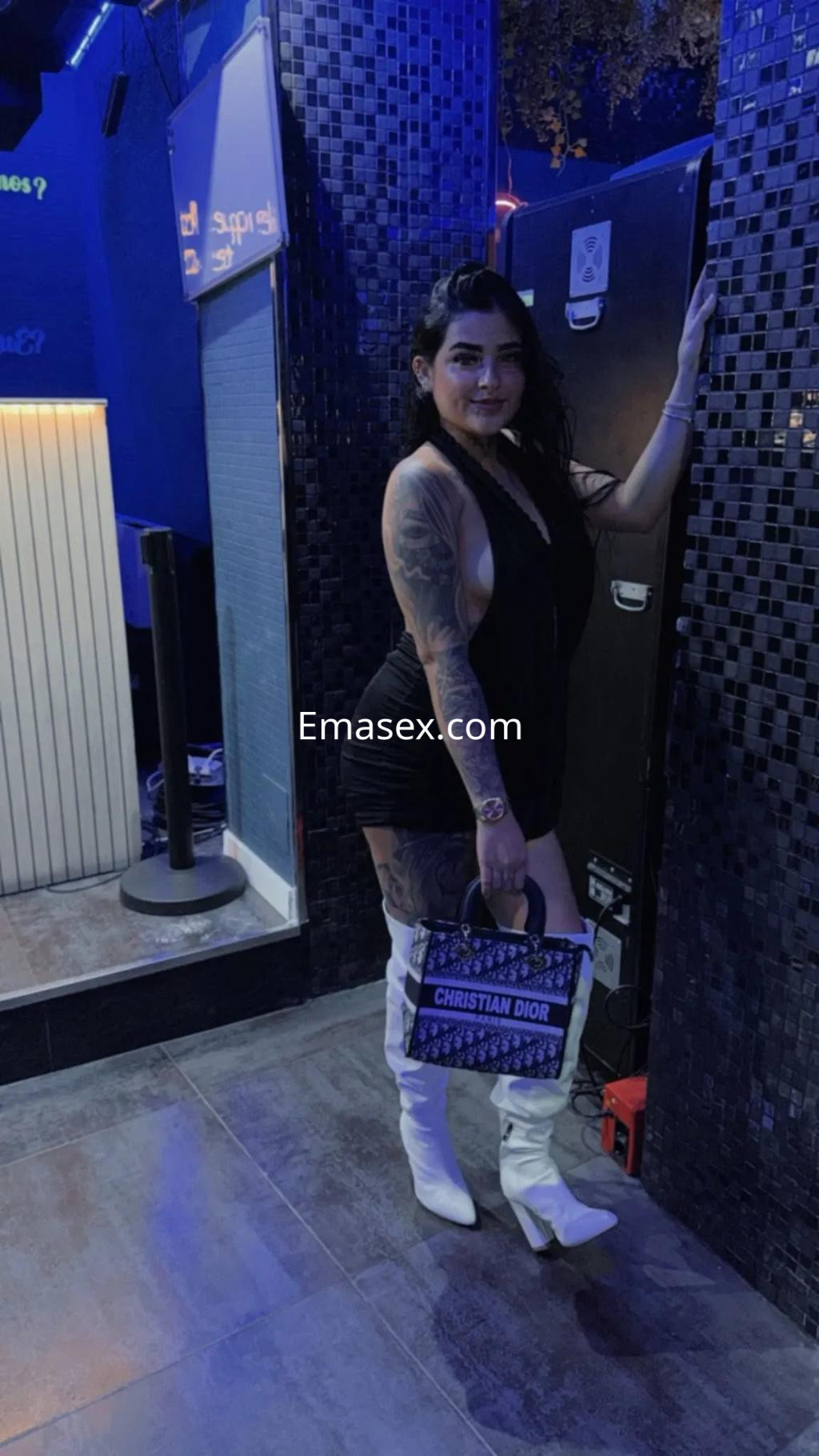Escort en Granada, con vestido negro ajustado y botas blancas, posando en un ambiente de luces azules.