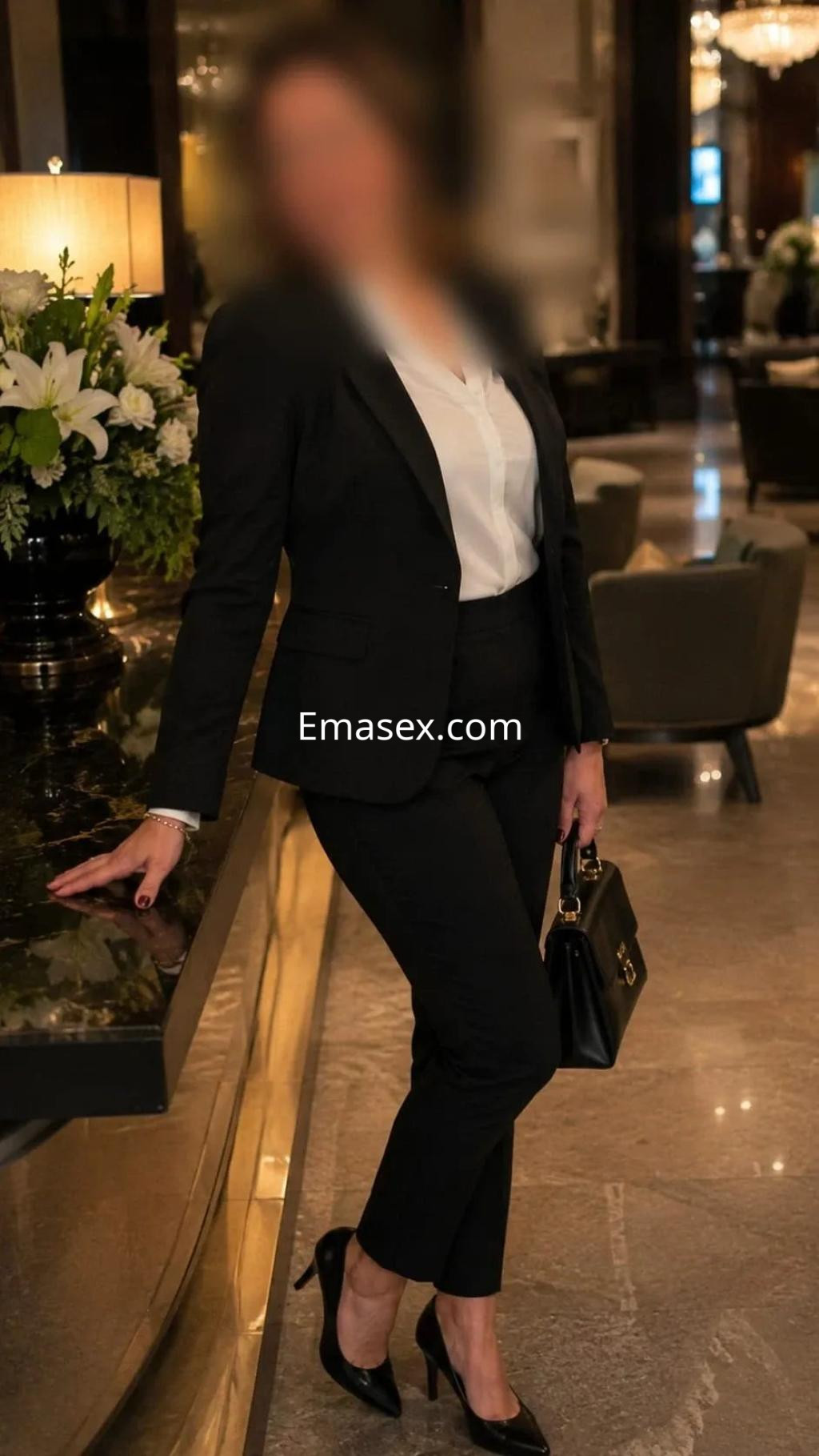 Puta en Madrid viste un elegante traje negro y tacones, posando con una bolsa de mano en un lujoso entorno.