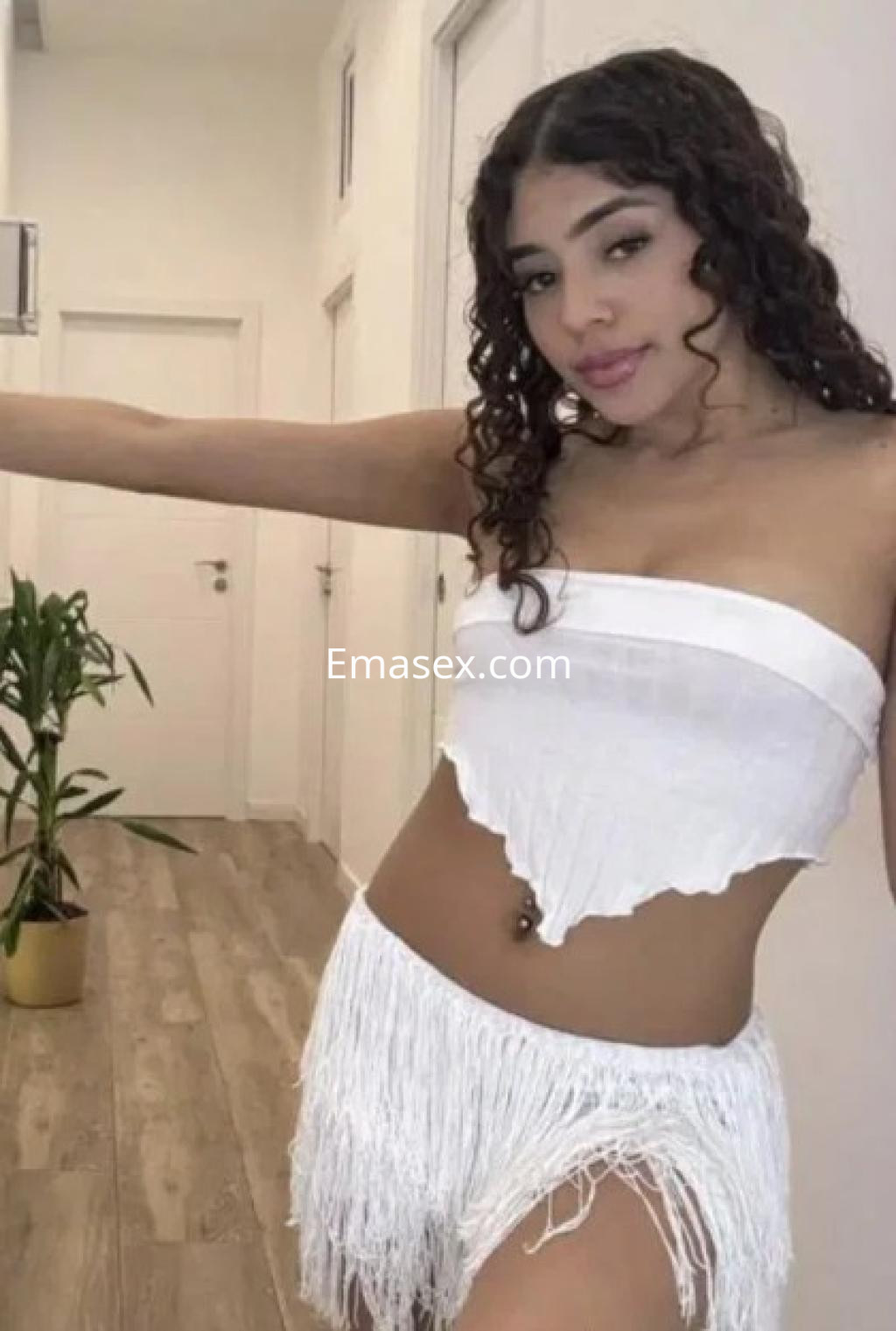 Escort en Málaga con top blanco sin mangas y pantalones cortos de flecos, posando en un pasillo iluminado.