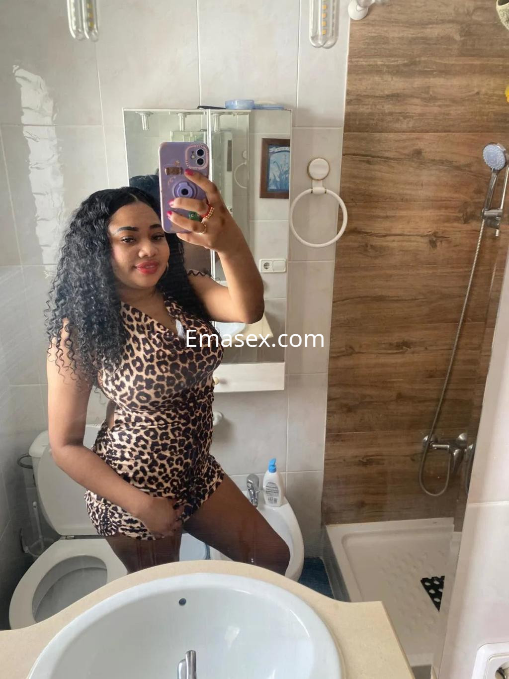 Escort en Sevilla, con cabello rizado y vestido de leopardo, posando en un baño moderno.