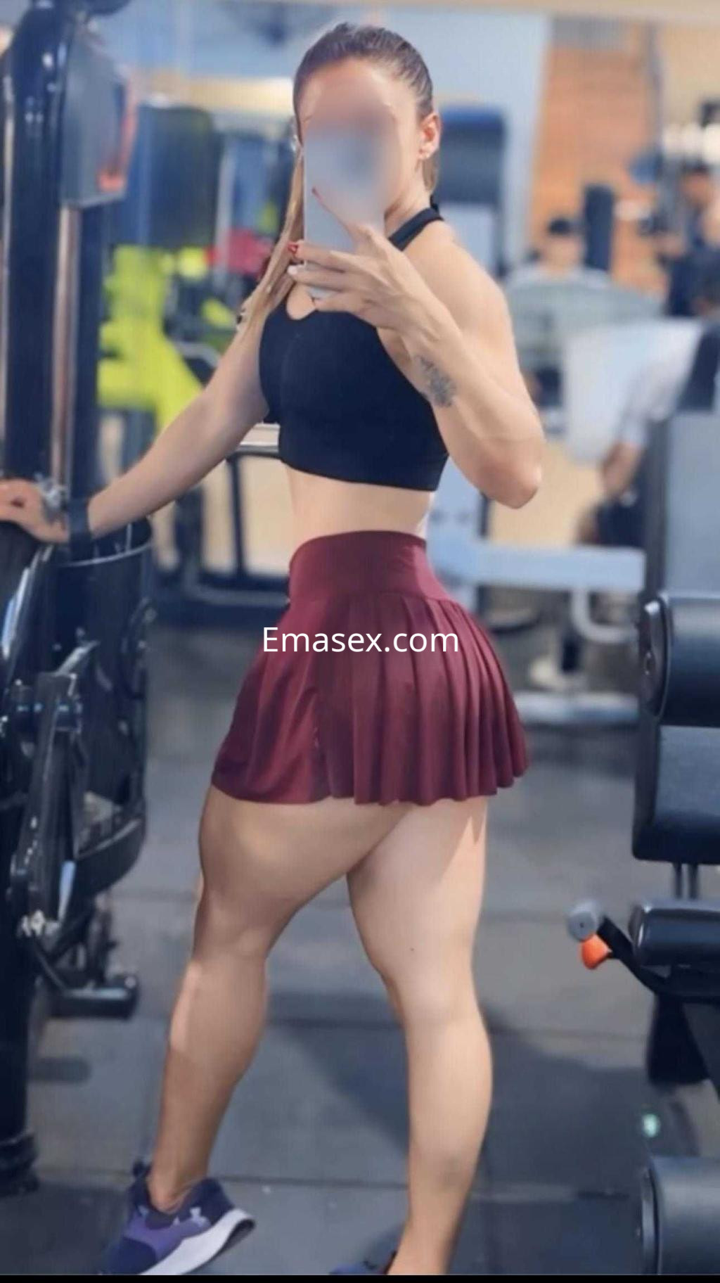La puta en Madrid viste un top negro y una falda corta roja, posando en un gimnasio con equipo de ejercicio.