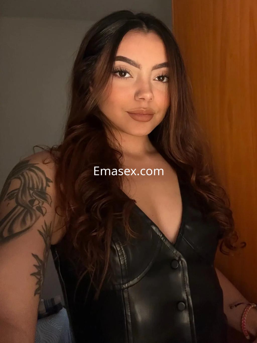 Escort en Alicante con cabello ondulado y suelto, viste un vestido de cuero negro y muestra un tatuaje en el brazo.
