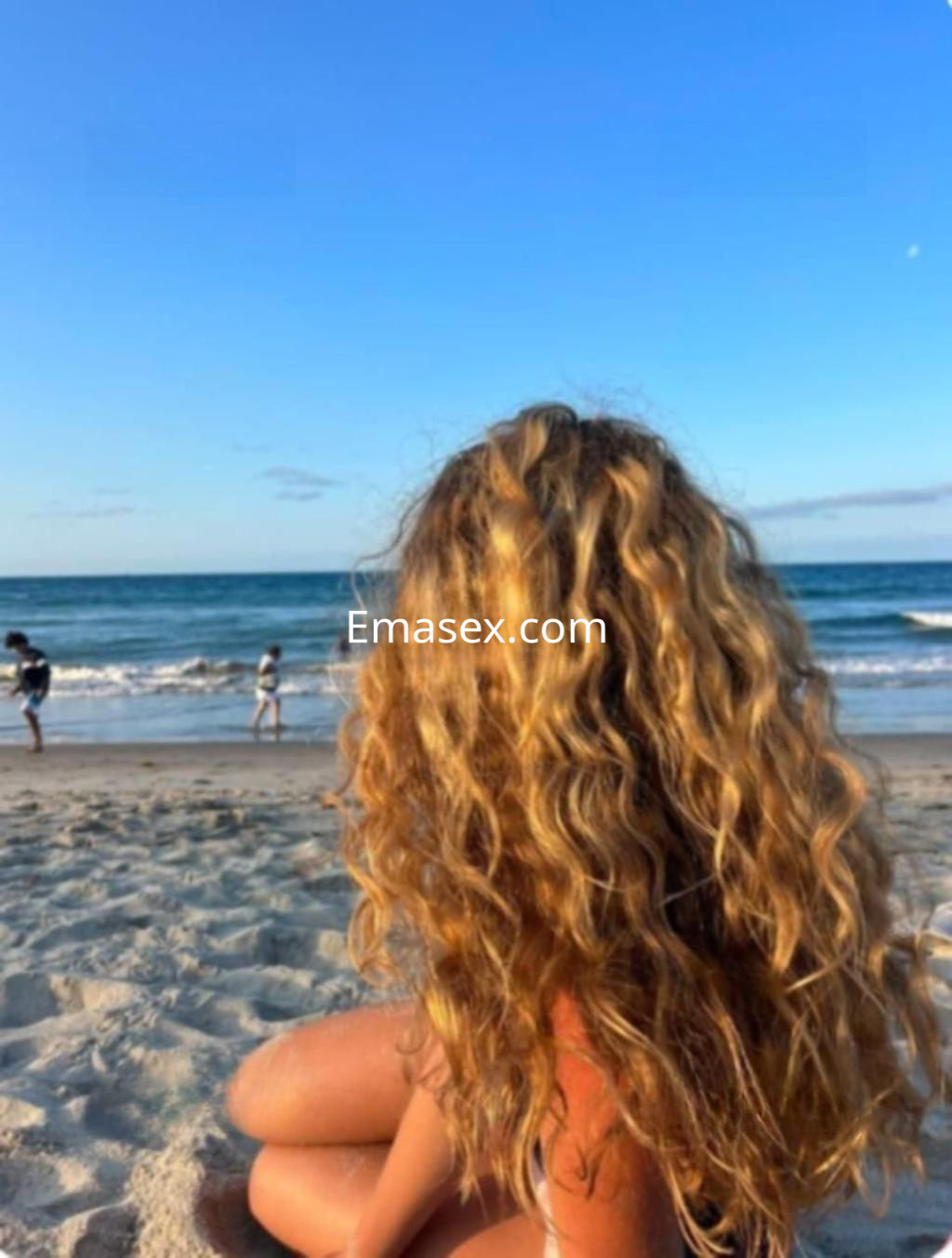 Escort con cabello rizado y dorado, sentada en la playa de Cádiz, mirando hacia el mar y el cielo despejado.