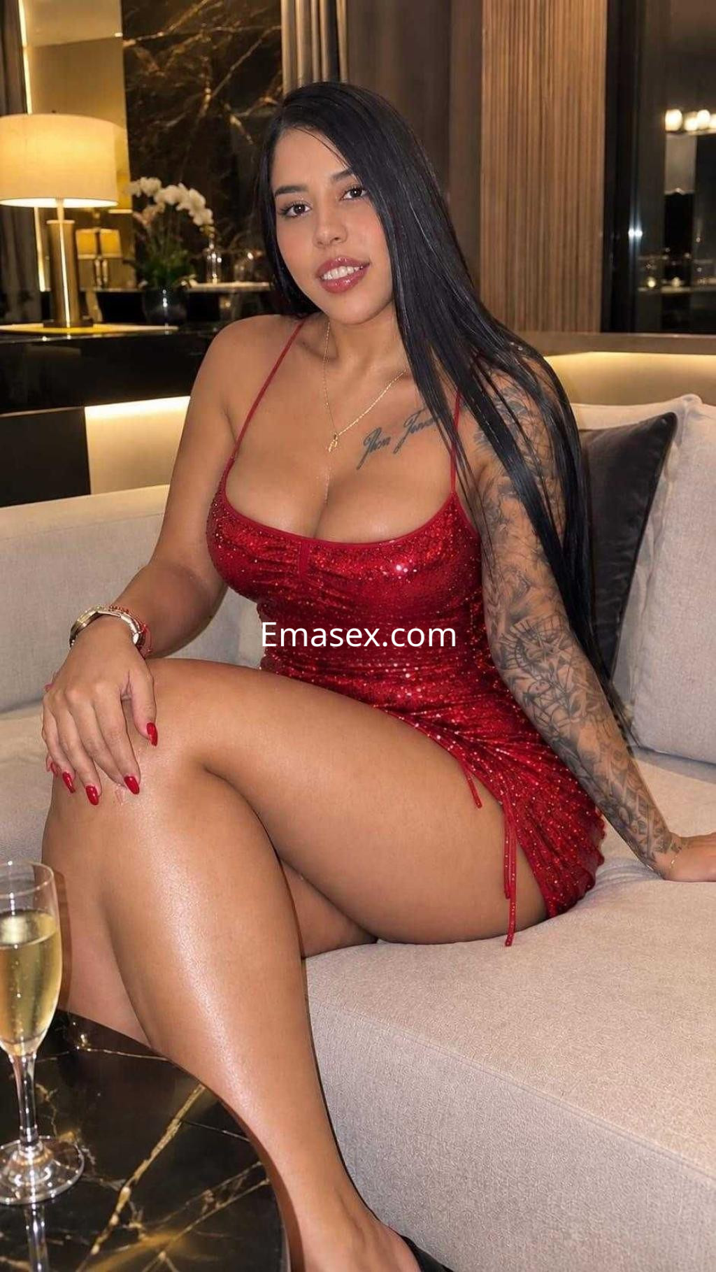 Puta en Granada, con vestido rojo brillante y cabello largo, posando sobre un sofá elegante en un ambiente sofisticado.