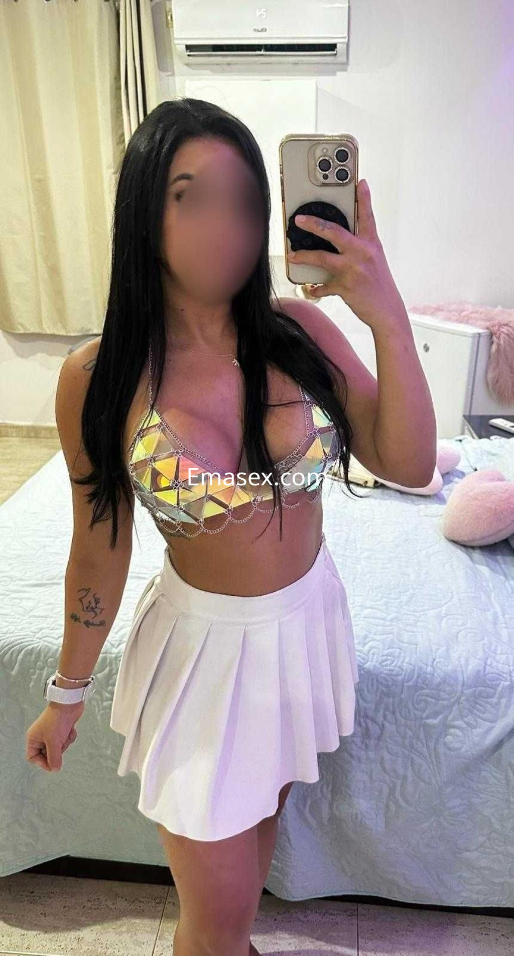 Escort en Madrid posando frente al espejo, vistiendo un sujetador brillante y una falda corta blanca.