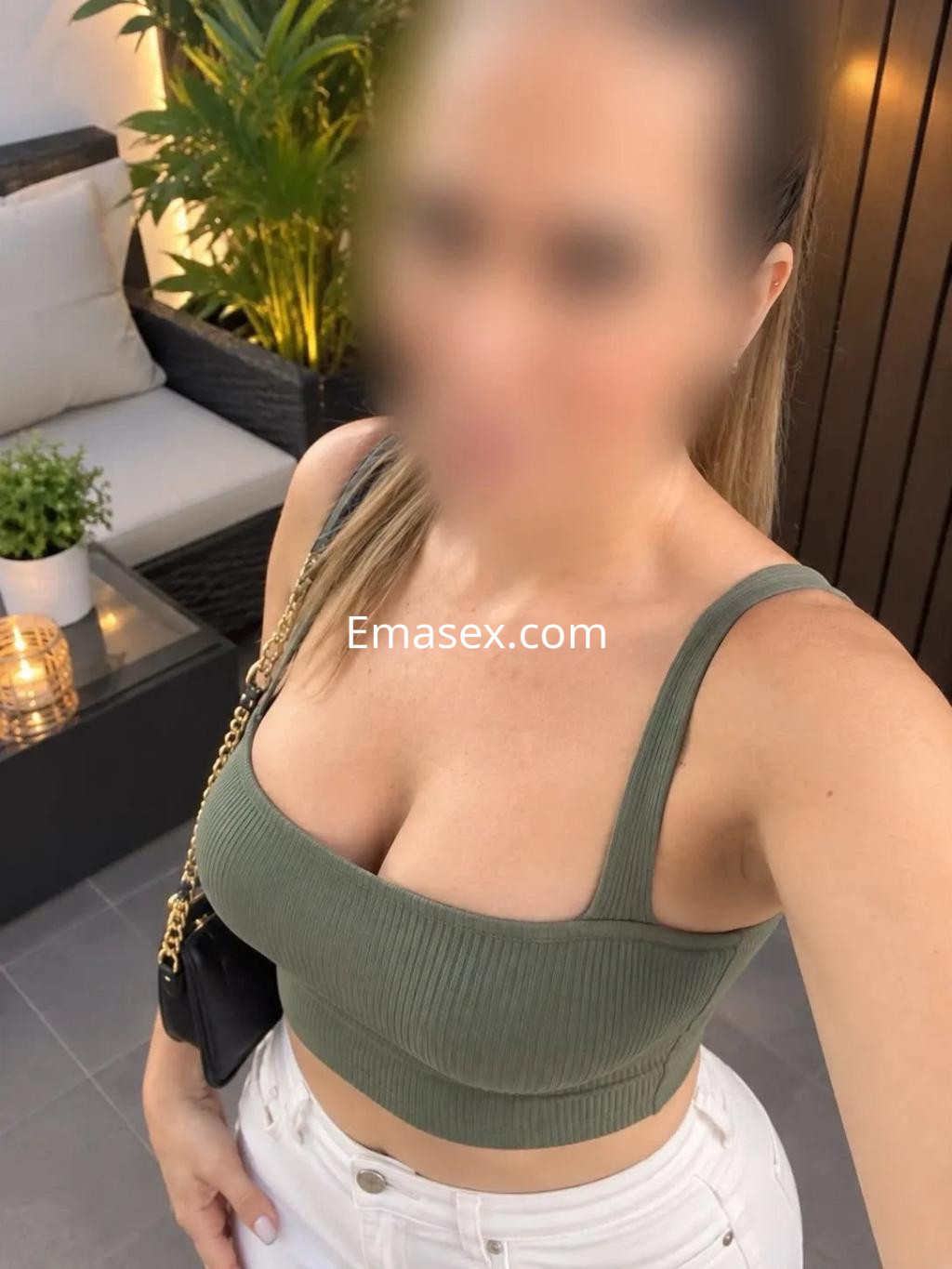 La escort en Valladolid luce un top verde ajustado y pantalones blancos, posando con un fondo elegante y plantas.