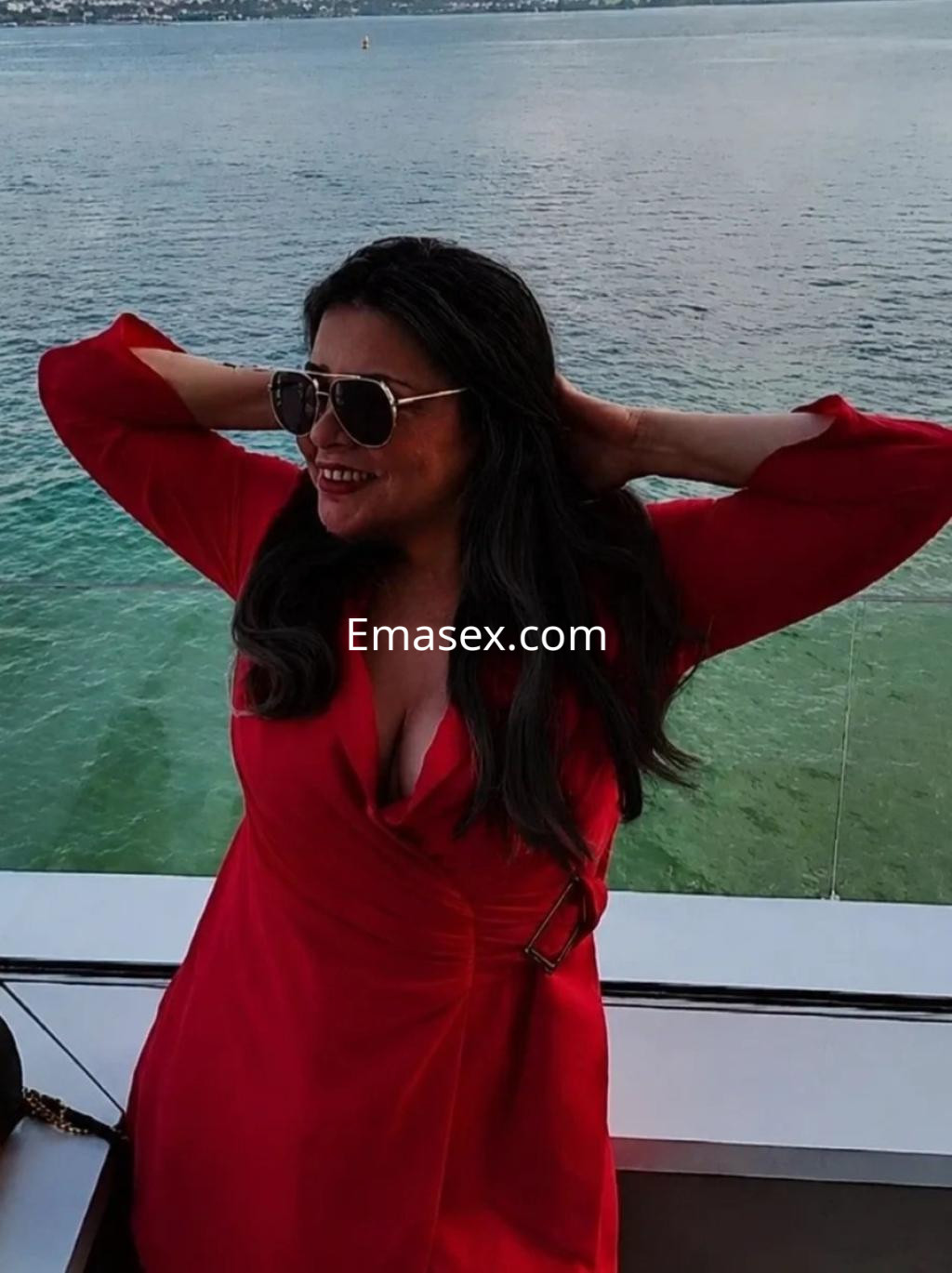 La escort en Santander luce un vestido rojo con escote, con gafas de sol y cabello largo y oscuro, frente al mar.