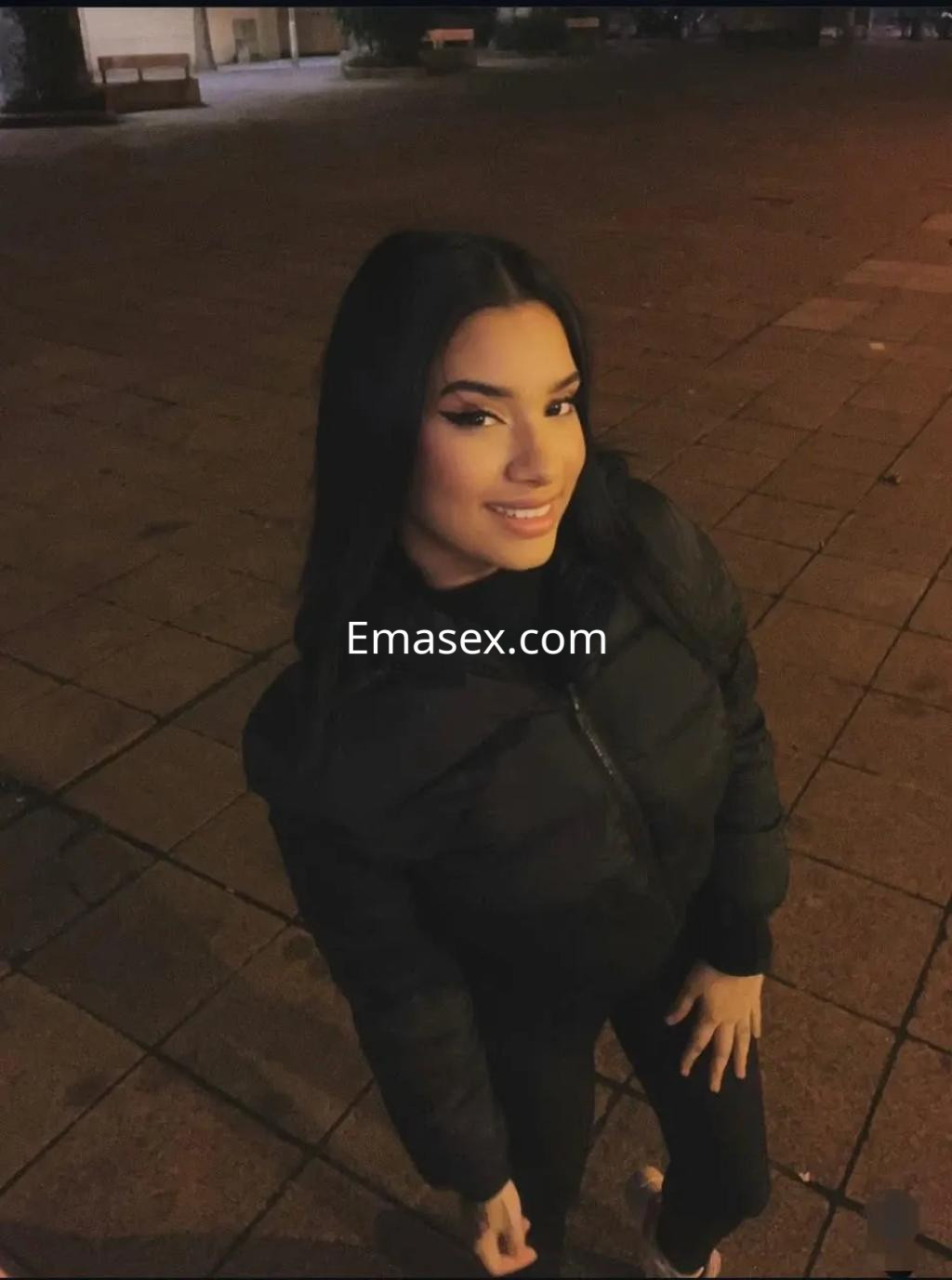 Escort en Granada, sonriendo con un abrigo negro, en un entorno urbano iluminado por la noche.