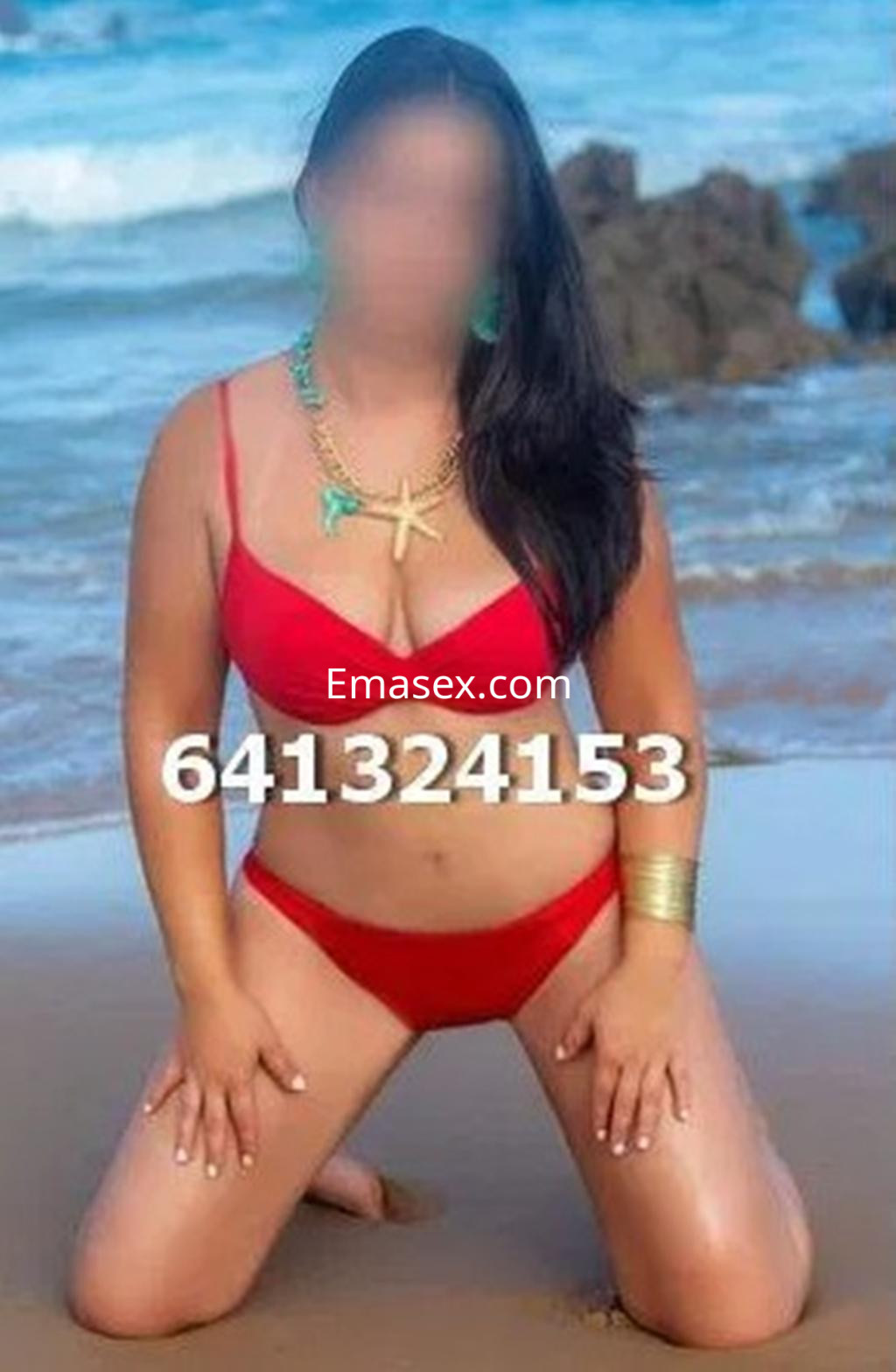 Escort en Burgos, posando de rodillas en la playa con bikini rojo y accesorios dorados, frente al mar.