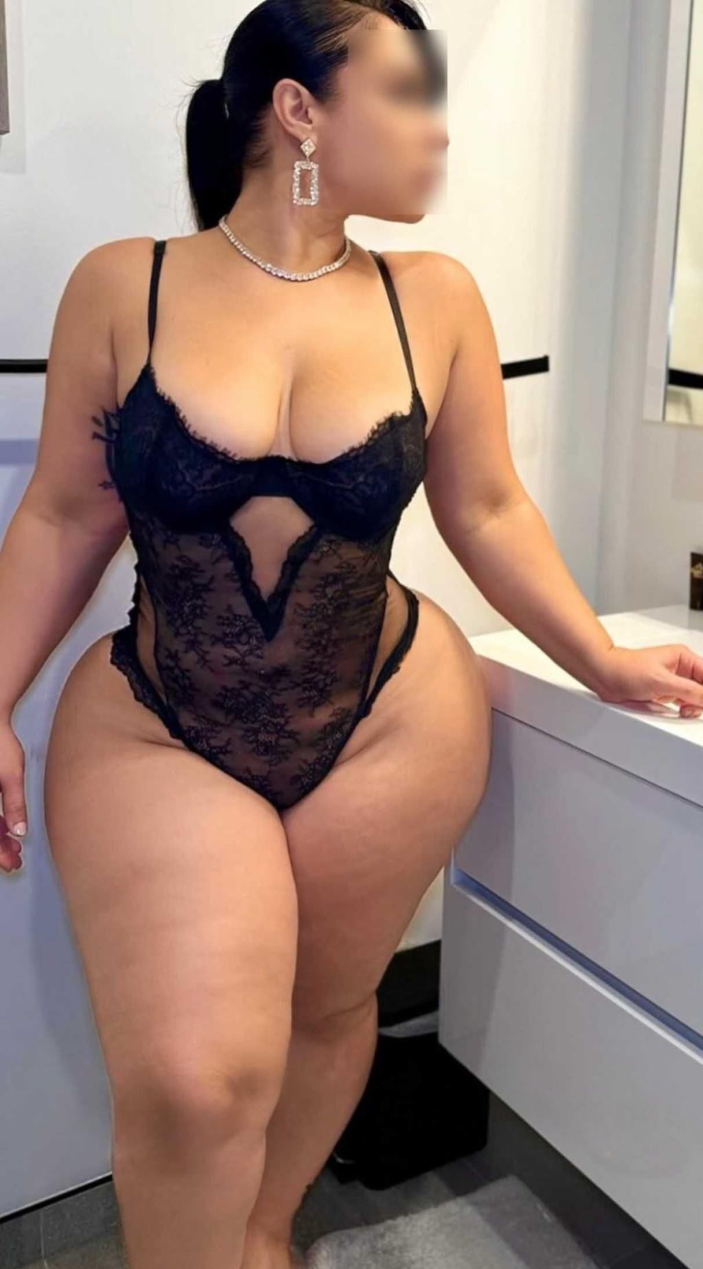 Escort en Madrid de pie junto a un mueble, luciendo un body negro de encaje y joyería, con cabello recogido.