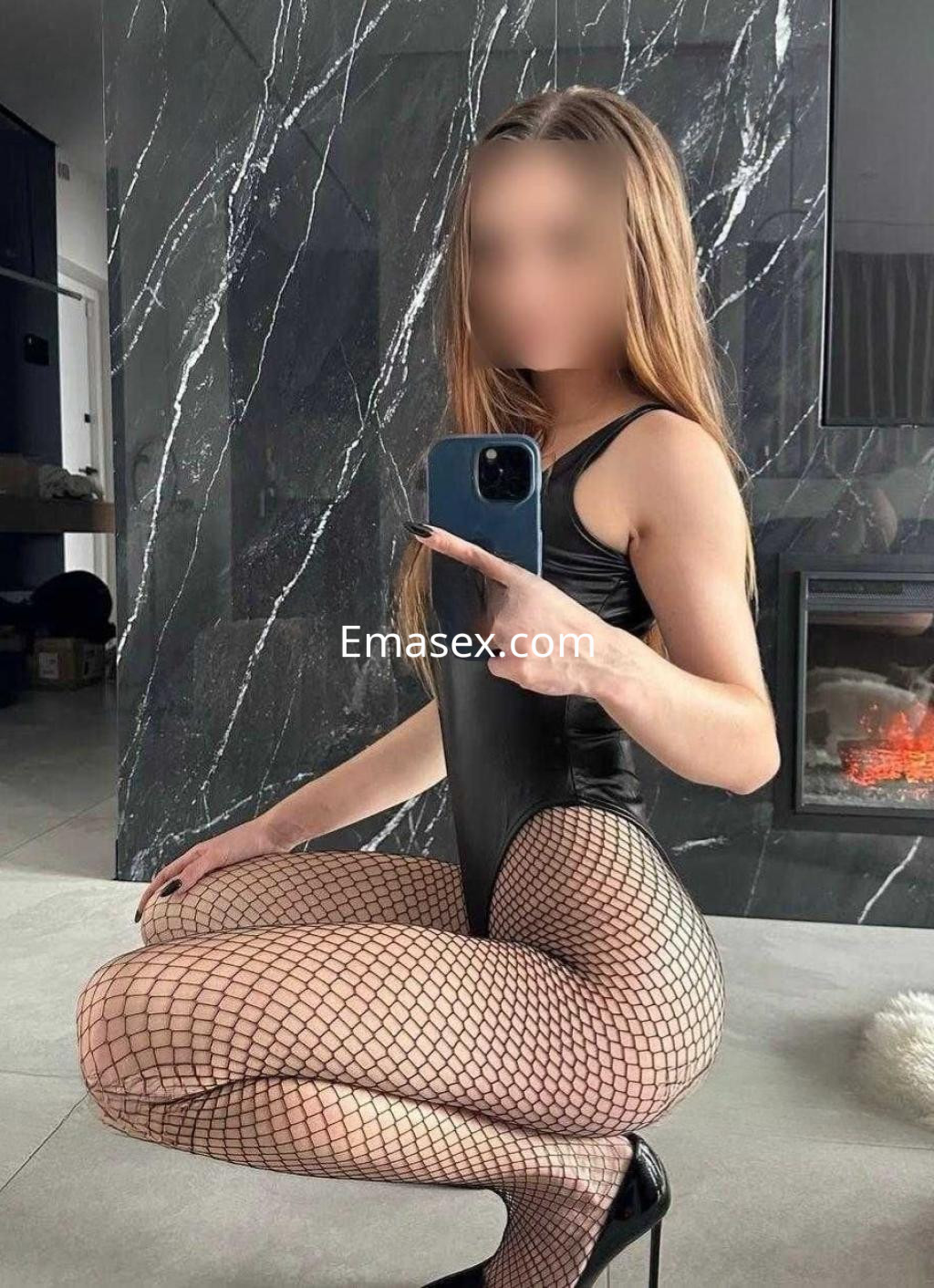 La puta en Madrid viste un body negro y mallas de rejilla, posando sentada con un teléfono en la mano.