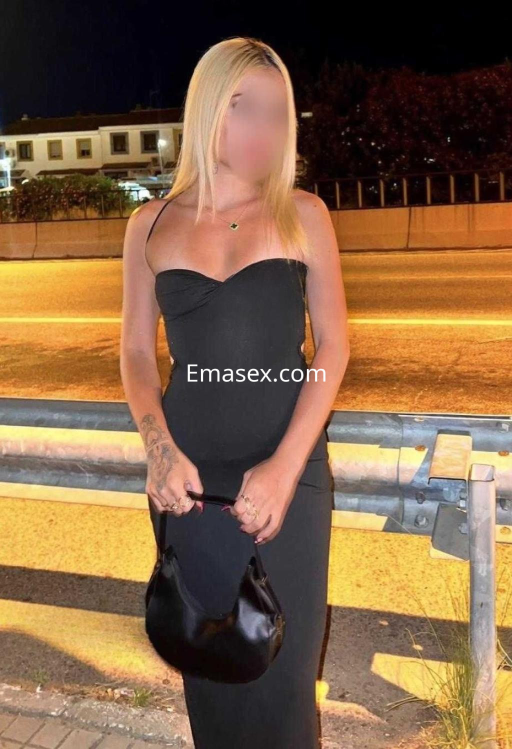 Escort en Madrid, vistiendo un vestido negro ajustado y sosteniendo un bolso, con cabello rubio y tatuajes visibles.