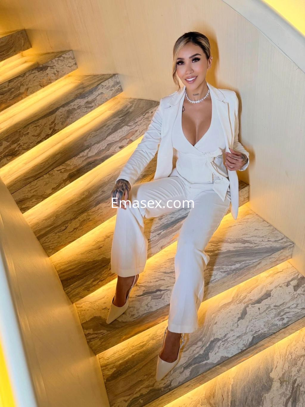 Escort con cabello rubio, vestida con un traje blanco, sentada en una escalera iluminada en Madrid.