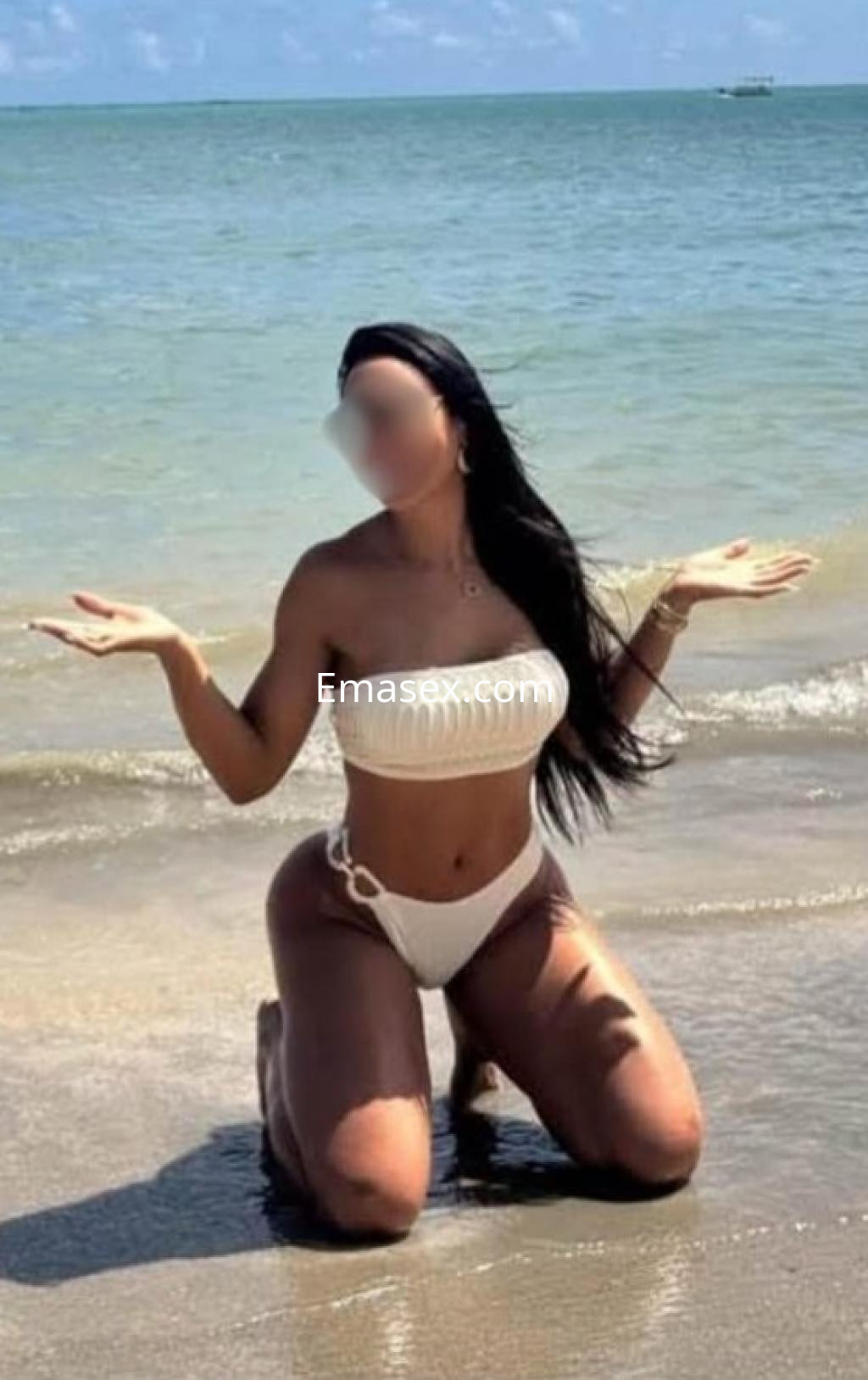 Escort en Bilbao de rodillas en la playa, vistiendo un bikini blanco y mostrando una pose relajada con el mar de fondo.