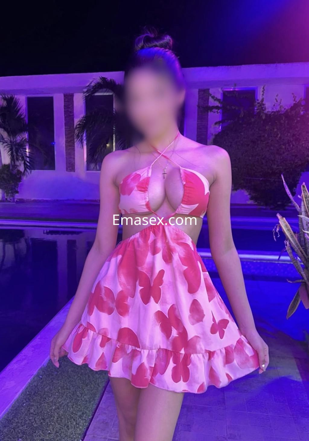 Puta de Donosti viste un vestido corto rosa con estampado de mariposas, posando junto a una piscina iluminada.