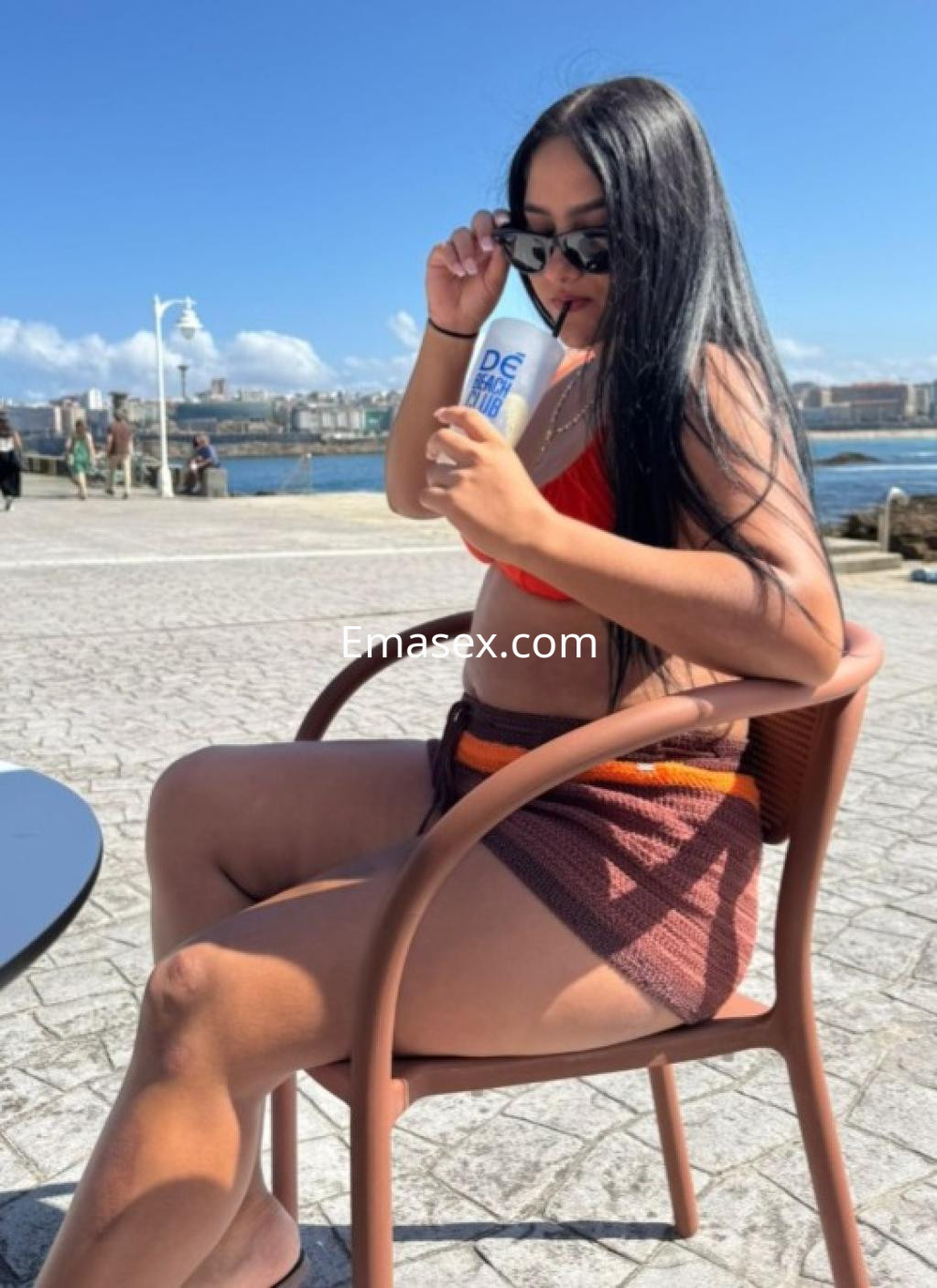 Puta en Coruña, sentada y tomando una bebida, viste un bikini rojo y un short marrón, con cabello largo y lacio.
