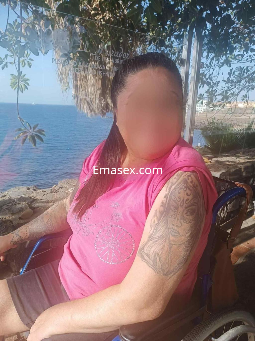 Escort en Tenerife sentada junto al mar, vistiendo una camiseta rosa y mostrando un tatuaje en el brazo.