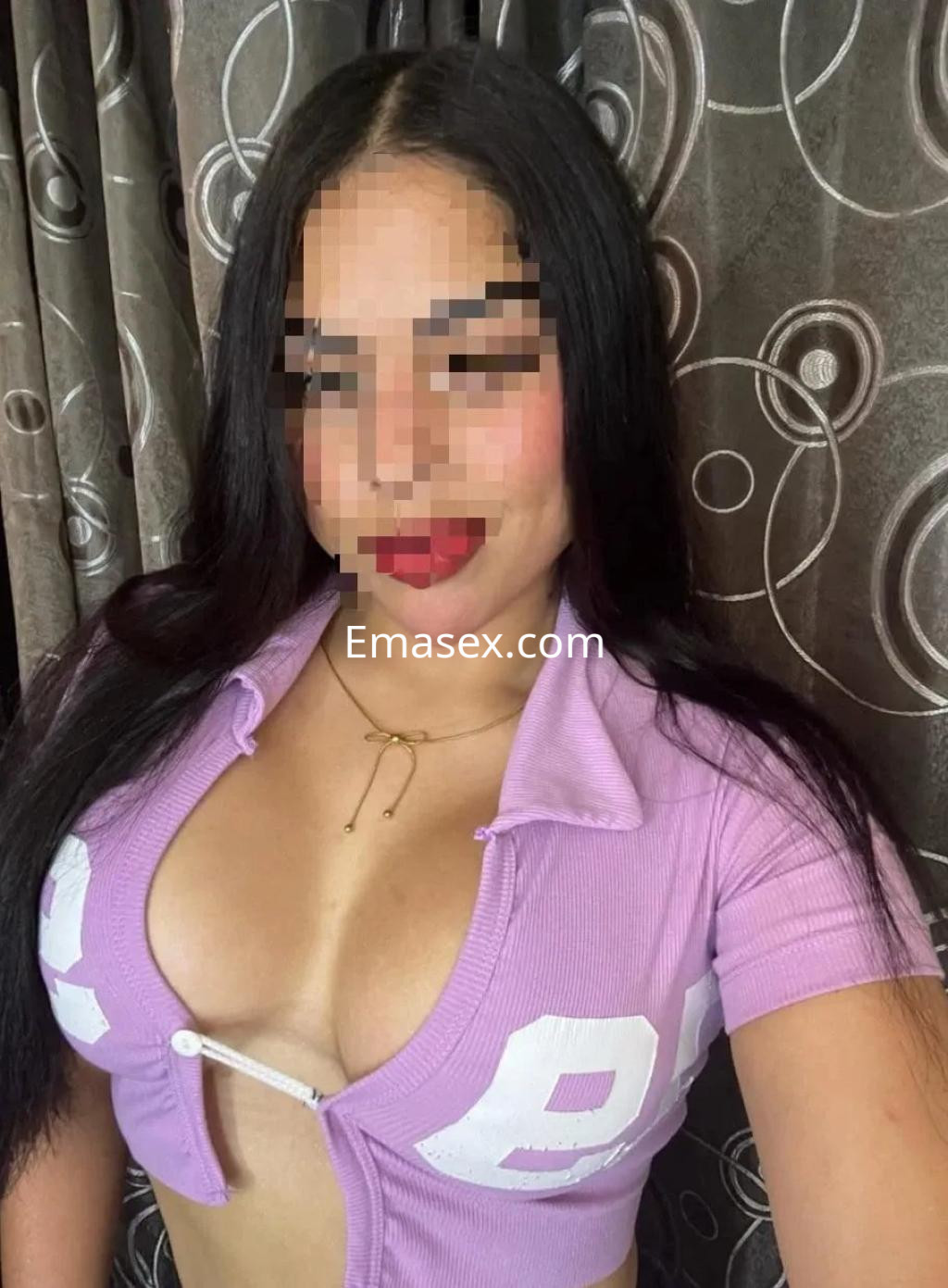 Escort en Alicante con cabello largo y oscuro, vistiendo una blusa rosa ajustada con escote en un entorno interior.