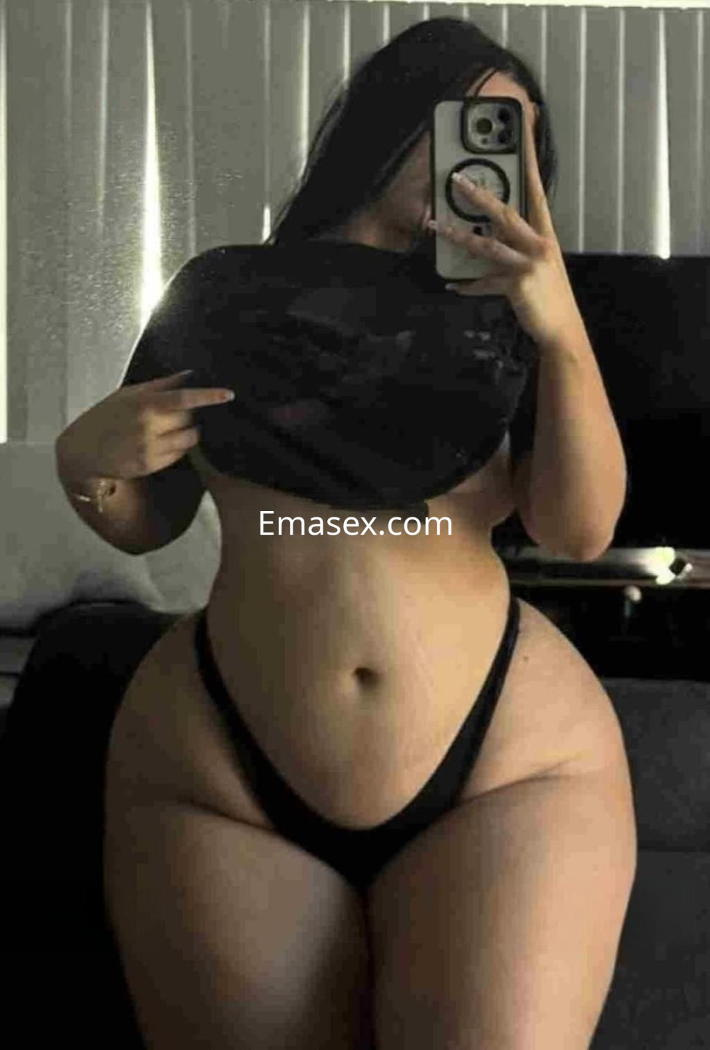 Puta en Sevilla, posando frente al espejo con una camiseta negra levantada y tanga negra, mostrando su figura.