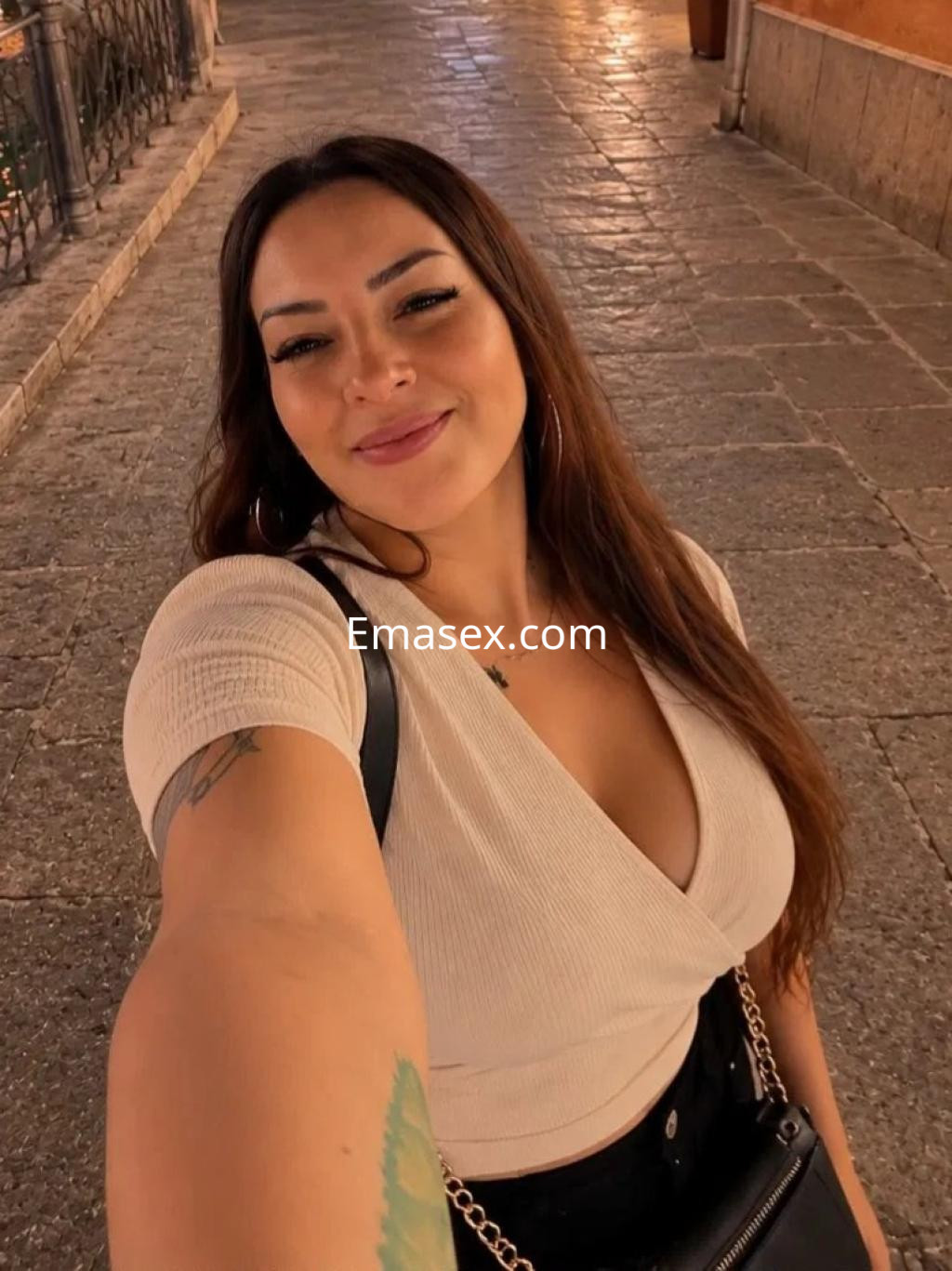Puta con cabello largo y ondulado, vistiendo un top claro y falda negra, sonriendo en una calle de Sevilla.