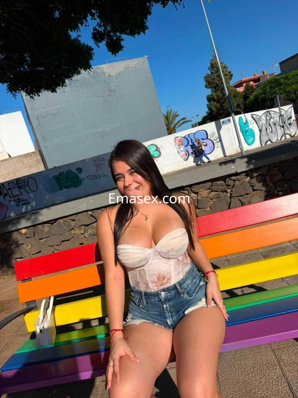 Escort en Tenerife, sentada en un banco multicolor con blusa ajustada y shorts de mezclilla, sonriendo al sol.
