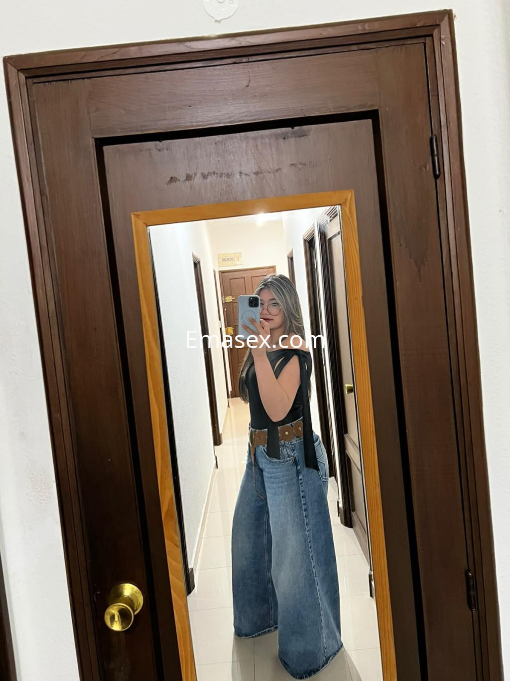 La puta se mira en un espejo, luciendo una blusa negra y unos pantalones vaqueros anchos en Valencia.