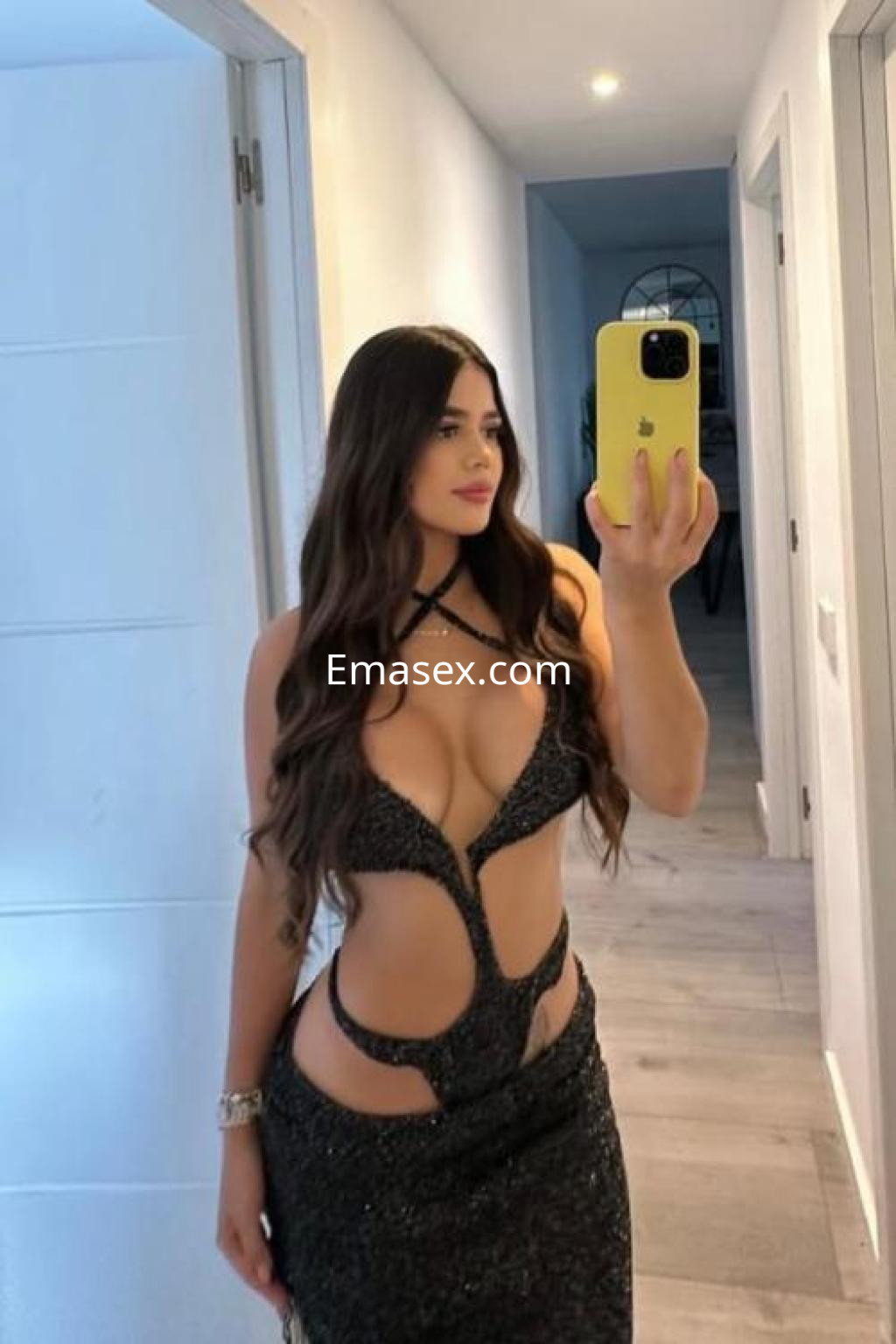Escort en Ibiza con un vestido negro brillante y ajustado, posando frente a un espejo con cabello largo.