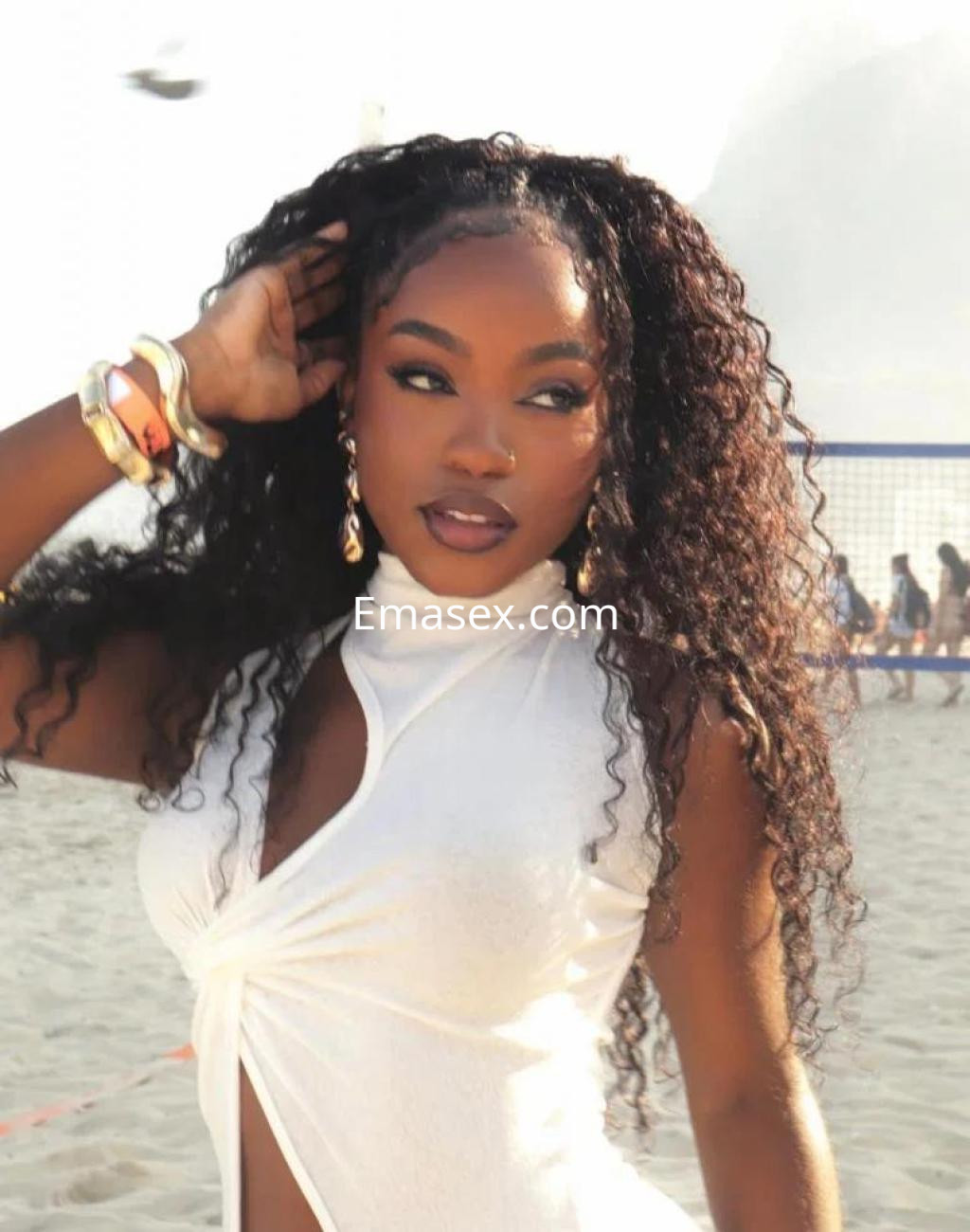 Escort en Alicante con cabello rizado y suelto, vestida con blusa blanca, posando en la playa con un fondo veraniego.