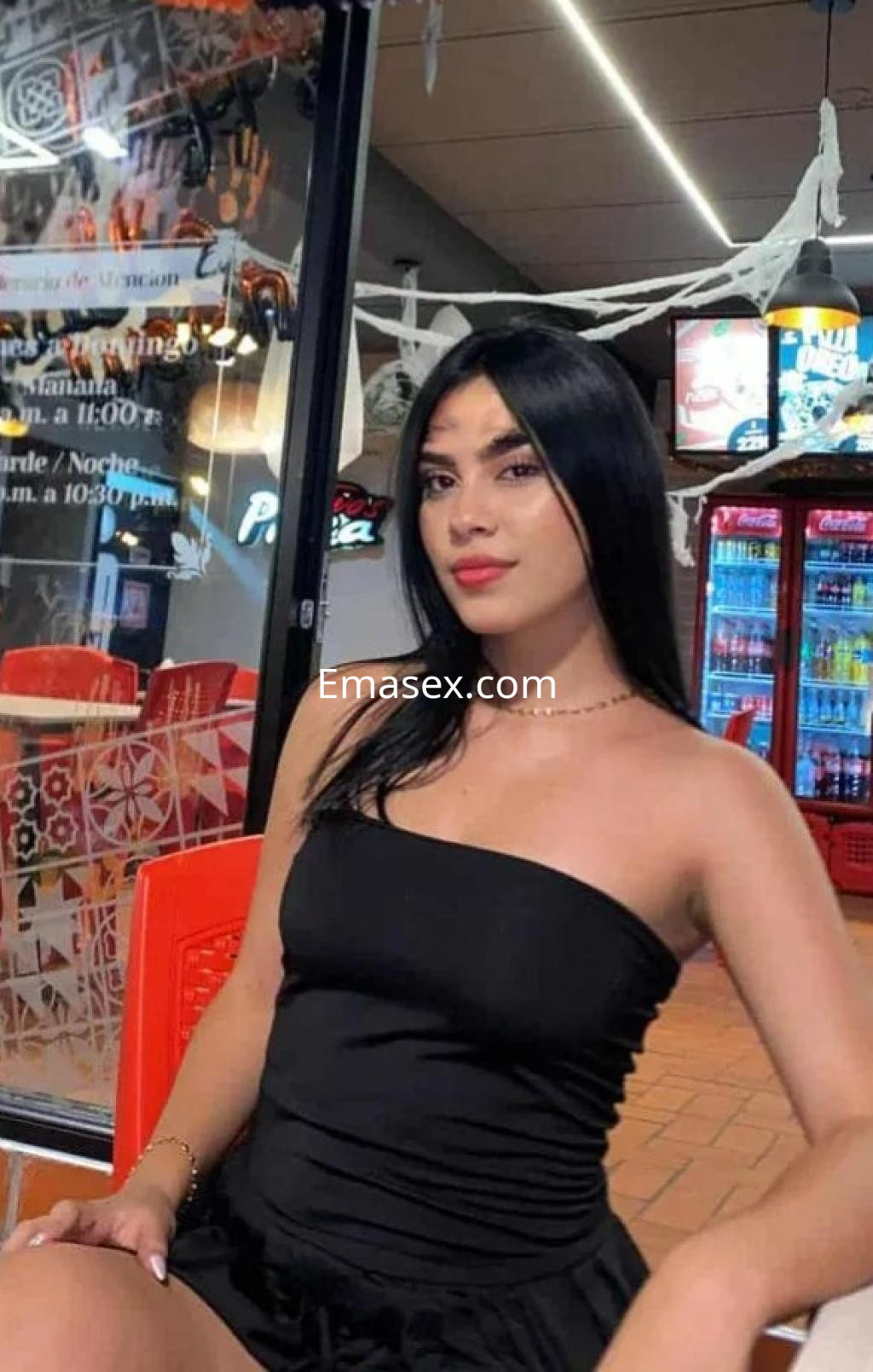 Escort en Gijón, con un vestido negro ajustado y cabello largo y lacio, posando en un local iluminado.