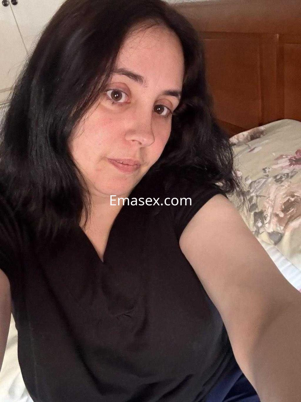 La puta en Barcelona muestra una expresión relajada mientras posa con una camiseta negra en su cama.