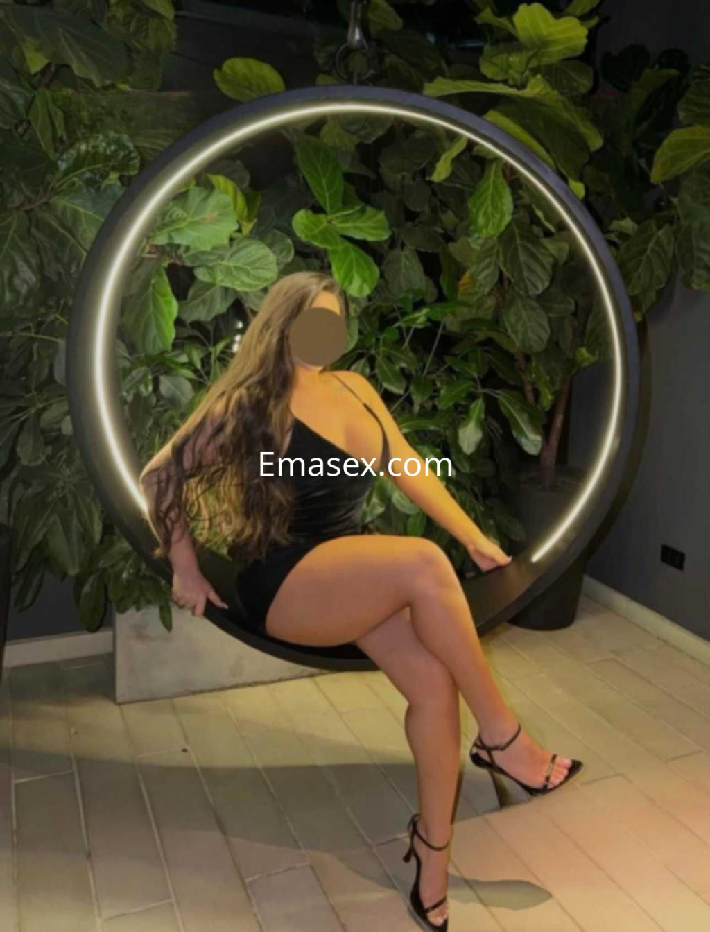 Escort en Toledo sentada en un columpio circular, vistiendo un vestido negro y tacones, rodeada de plantas.