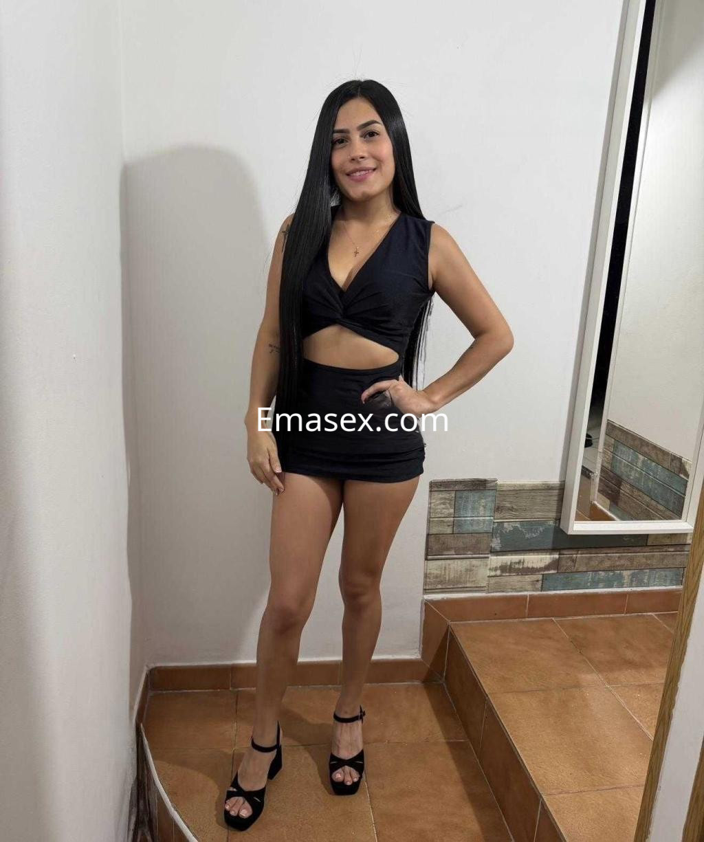 Escort en Madrid, luciendo un vestido negro ajustado y tacones, posando con una sonrisa en un entorno interior.
