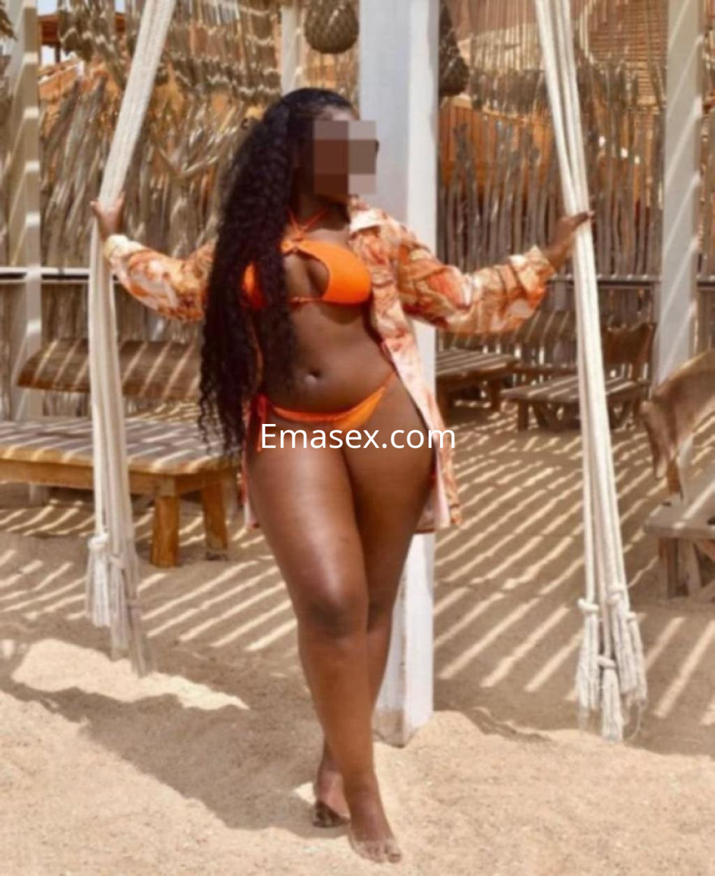 Puta en Cádiz posando en un bikini naranja con una blusa transparente, rodeada de un ambiente tropical.