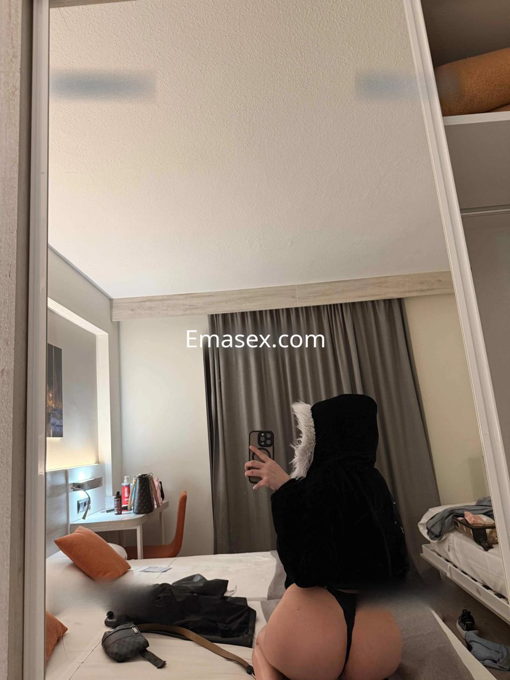 Escort en Granada se toma una selfie en el espejo, vistiendo un abrigo negro con capucha y lencería minimalista.