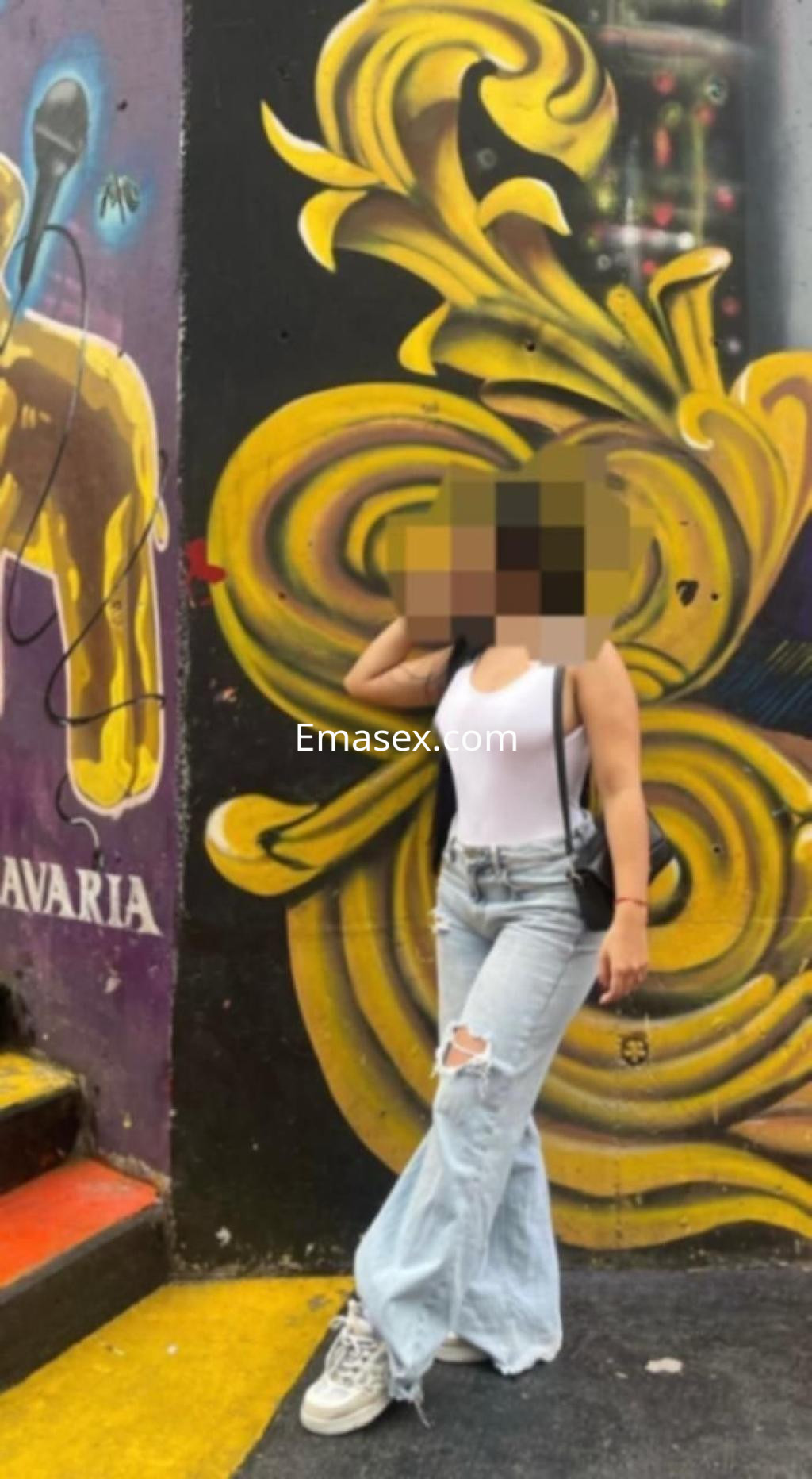 Escort luciendo una camiseta blanca y jeans rasgados, posando frente a un mural colorido en Toledo.