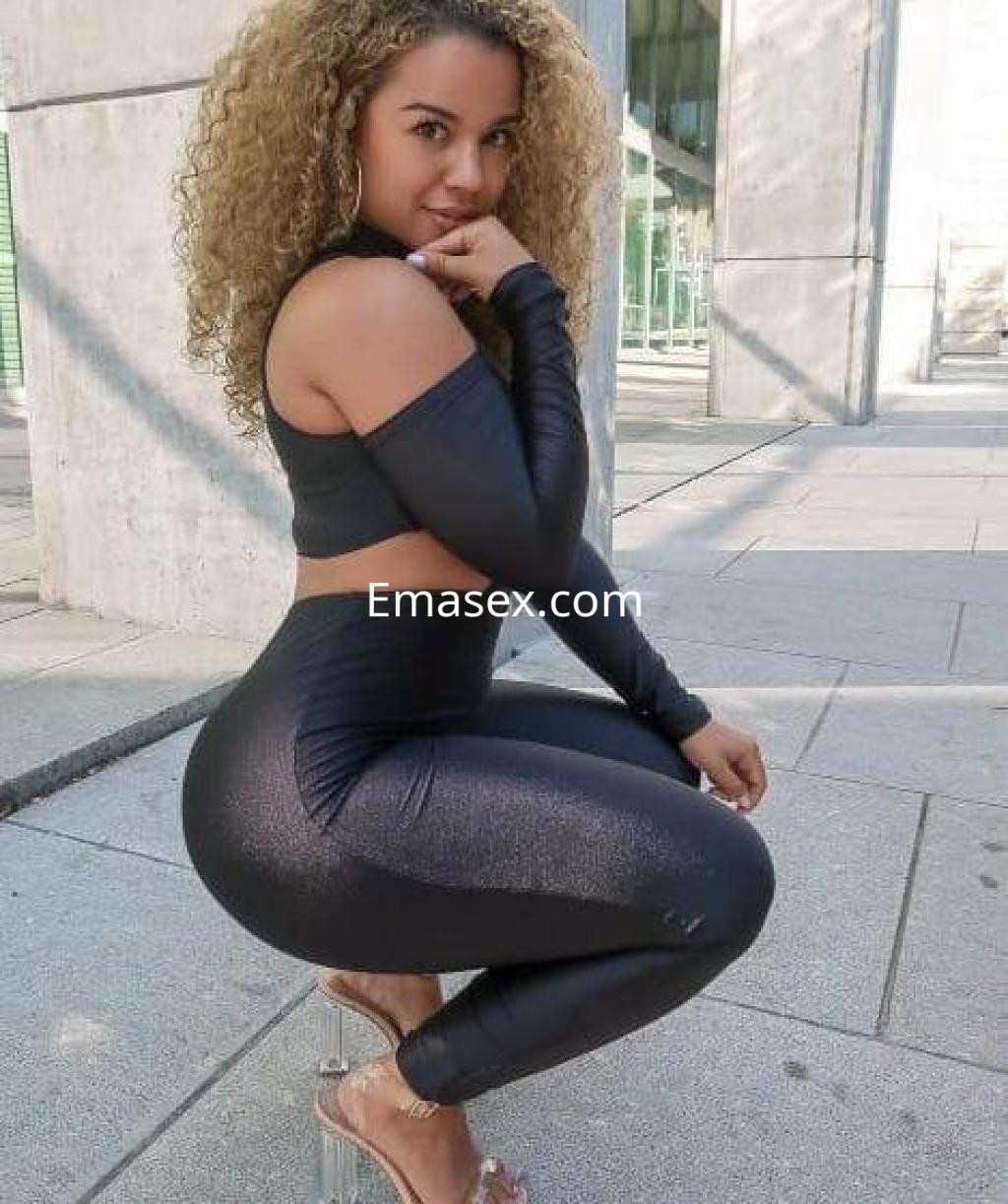 Escort posando en Granada con un conjunto negro ajustado, cabello rizado y tacones transparentes.