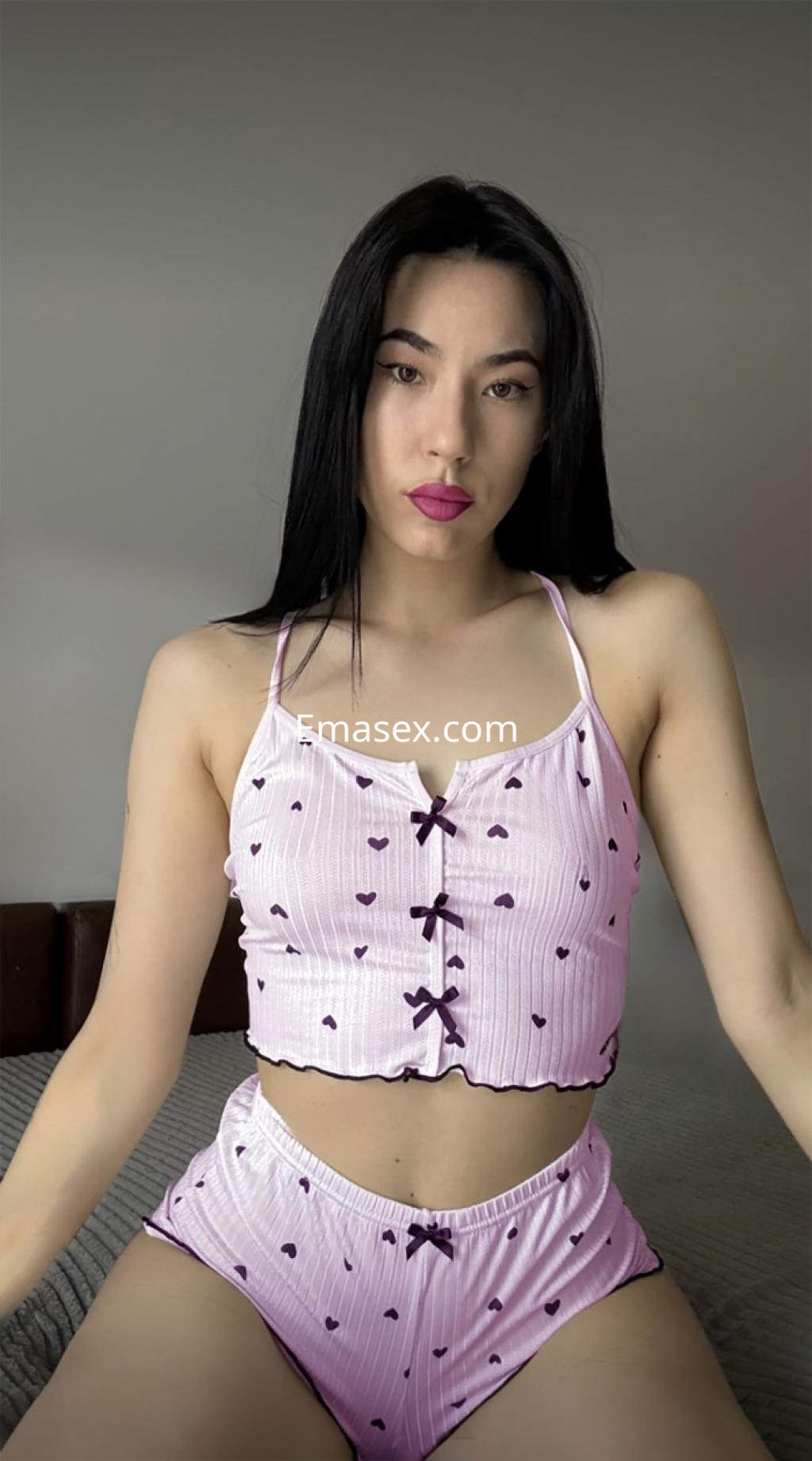 Puta en Tarragona viste un conjunto de lencería rosa con corazones, posando sentada en la cama.
