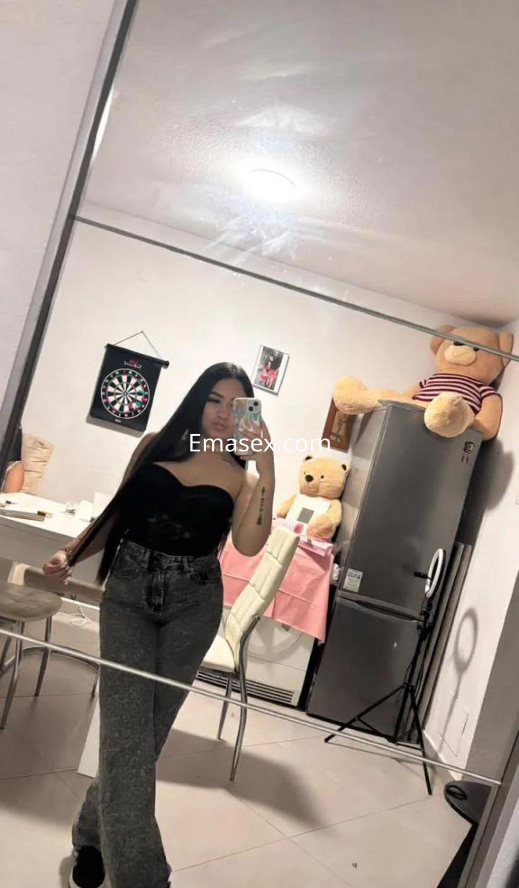 Escort en Salamanca con cabello largo y lacio, viste un top negro y jeans grises en un ambiente doméstico.