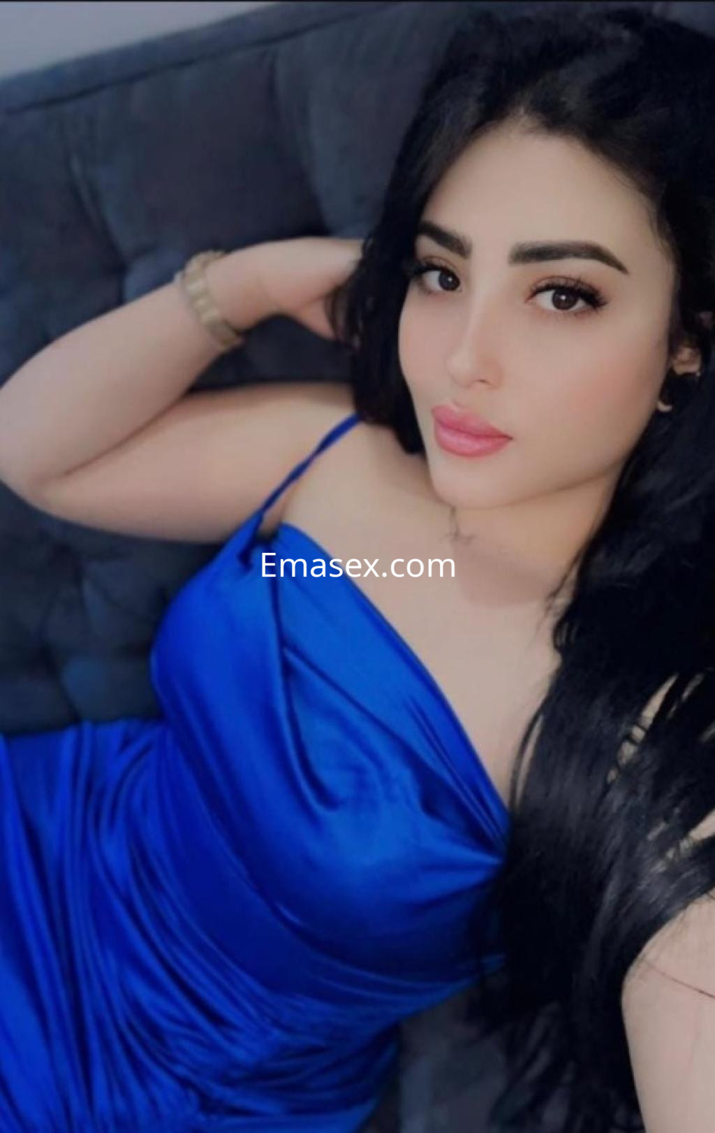 Puta posando sobre un sofá gris, luce un vestido azul ajustado y tiene el cabello largo y negro en Tarragona.