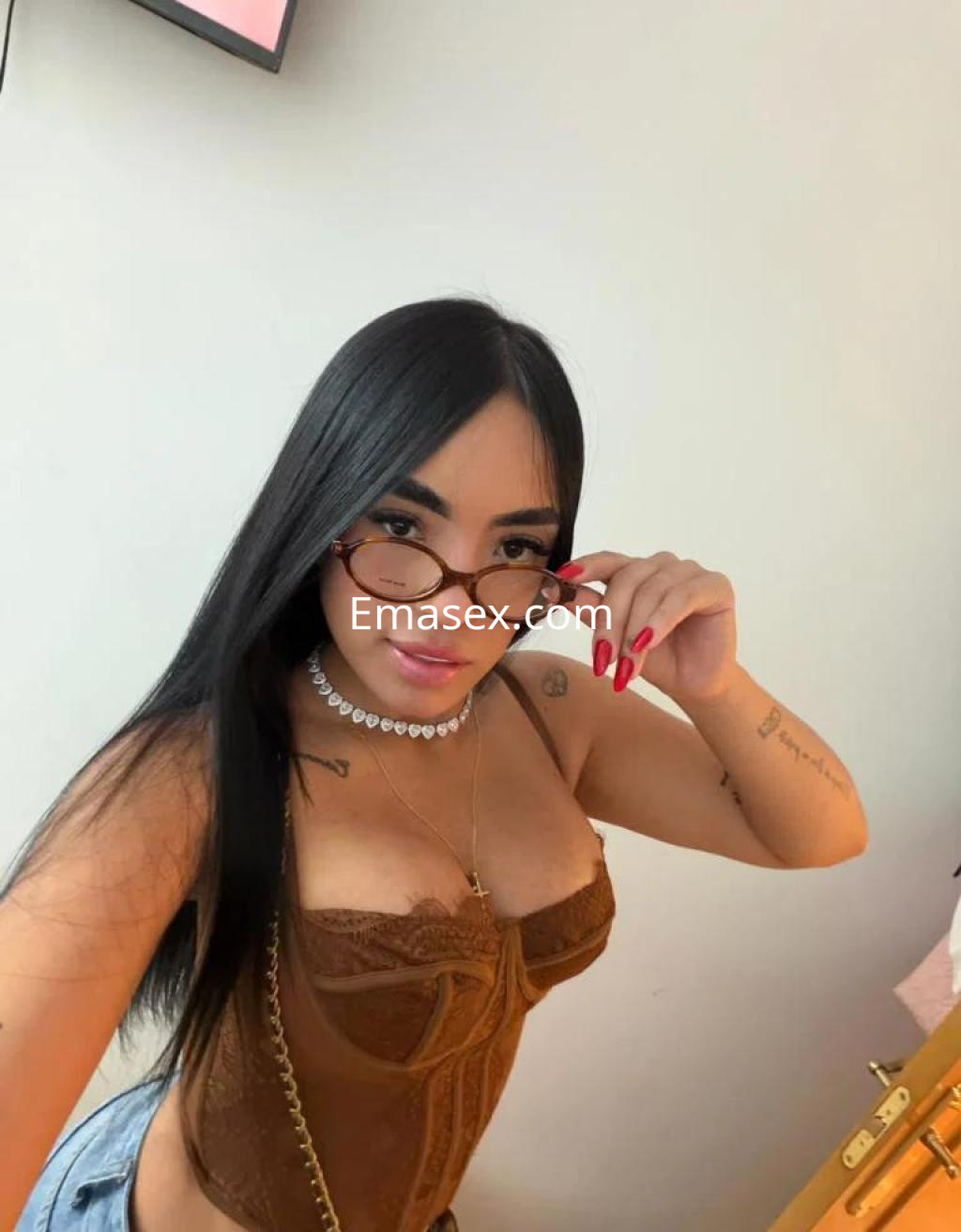 Escort en Toledo con cabello largo y oscuro, usando un top marrón y gafas, posando con una mano en la cara.