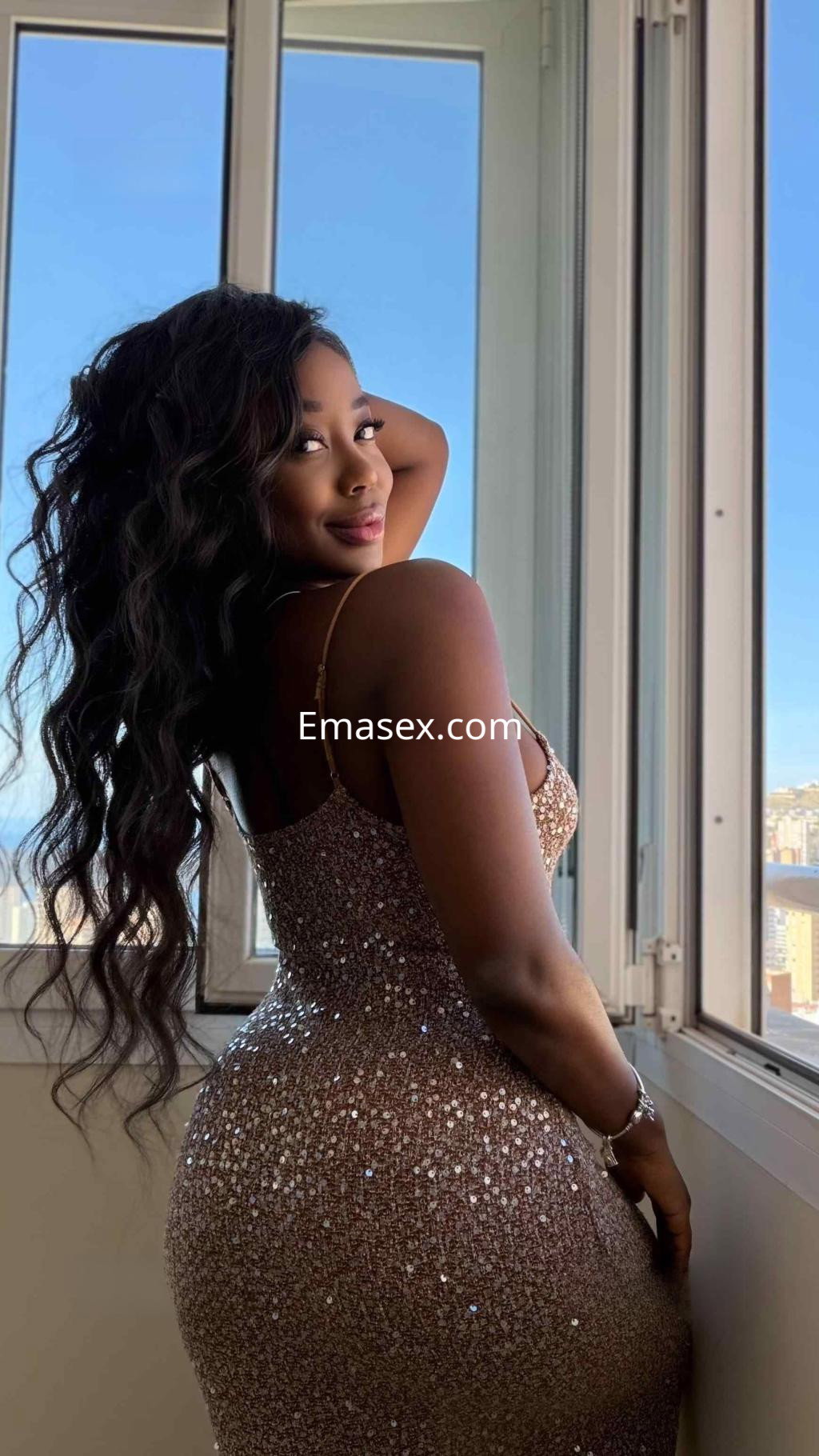Puta en Alicante con cabello rizado, viste un vestido brillante que acentúa su figura en un entorno iluminado.