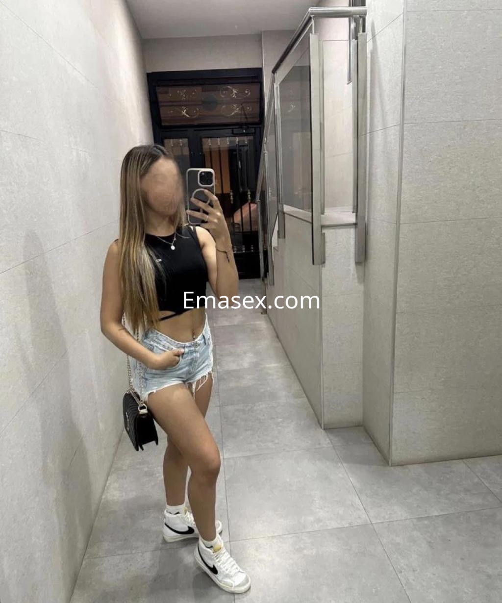 Puta en Toledo posa en un espejo, vistiendo un top negro y shorts de mezclilla, con zapatillas blancas.