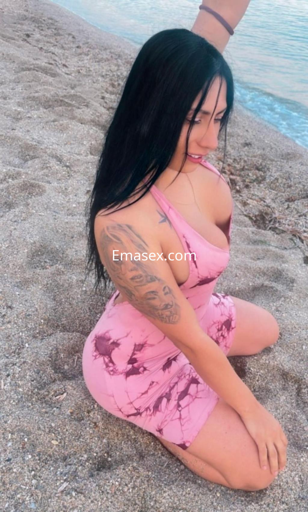 Escort en Gijón posando en la playa, con vestido rosa ajustado y cabello largo, mostrando un tatuaje en su brazo.